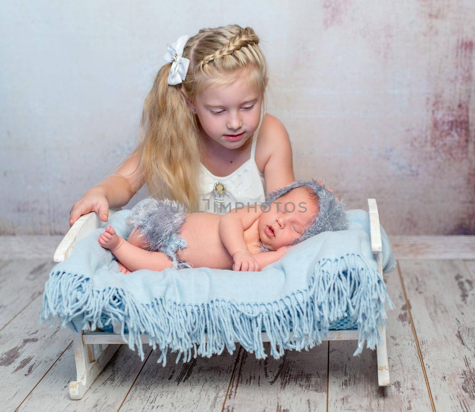 sleeping newborn boy on little bed with blue diaper and older sister watching him beside the bed