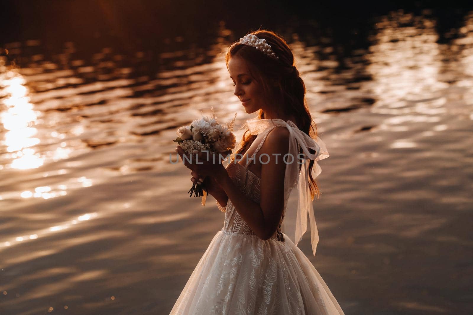 An elegant bride in a white dress and gloves stands by the river in the Park with a bouquet, enjoying nature at sunset.A model in a wedding dress and gloves in a nature Park.Belarus by Lobachad