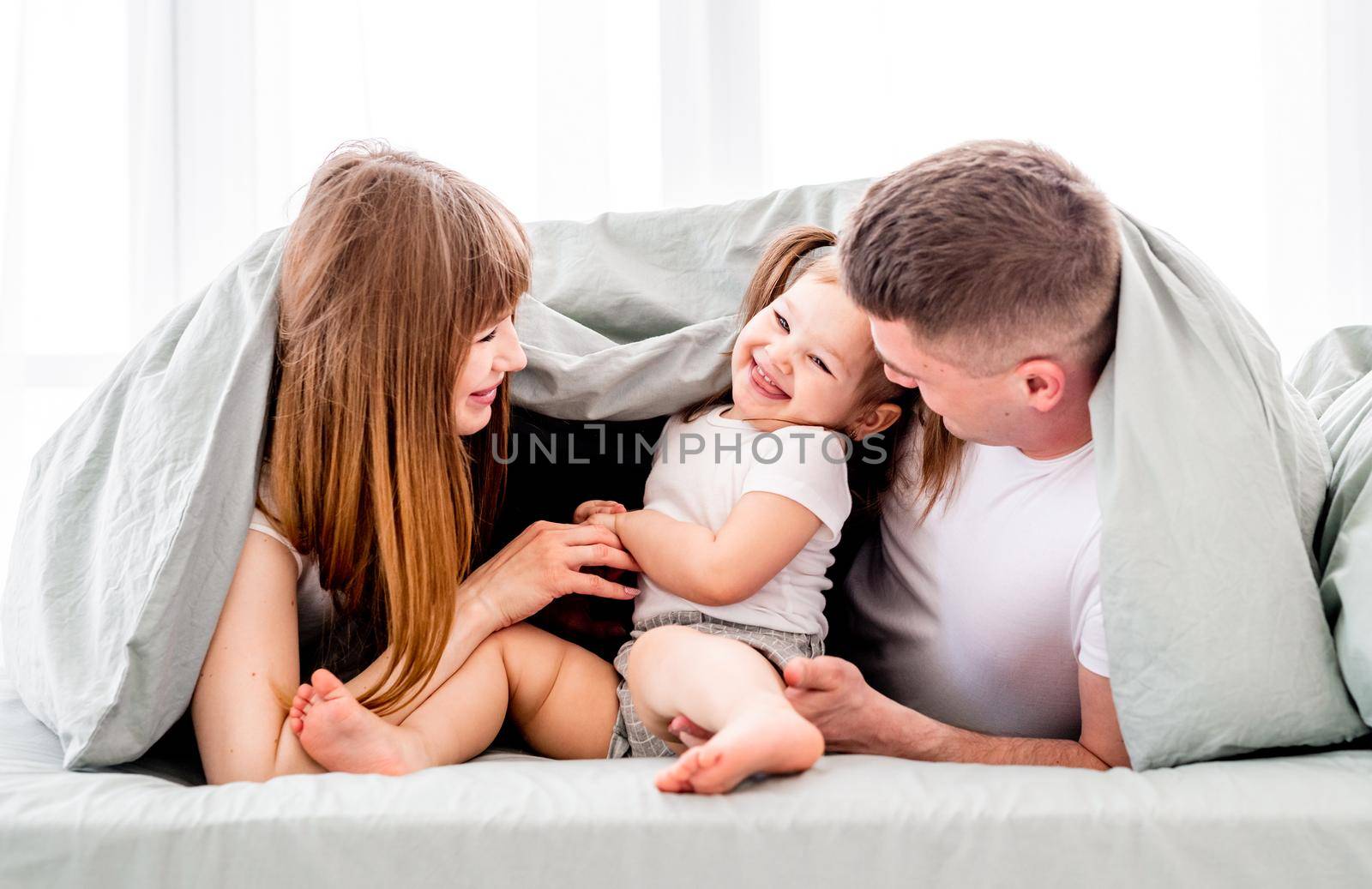 Family under the blanket in the bed. Young parents playing with small daughter under coverlet and smiling. Beautiful girl with her husband and kid in the bedroom