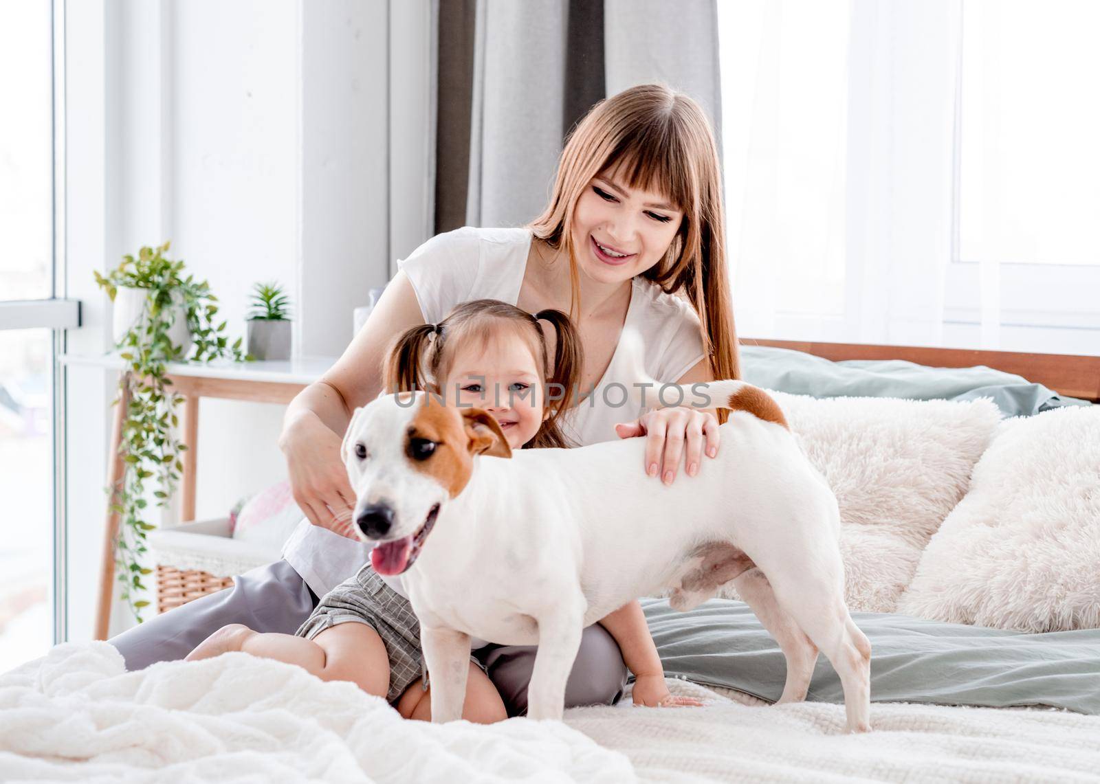 Beautiful young mother sitting in the bed with little daughter, petting cute dog and smiling. Attractive girl mom with her child and pet in the bedroom. Family portrait
