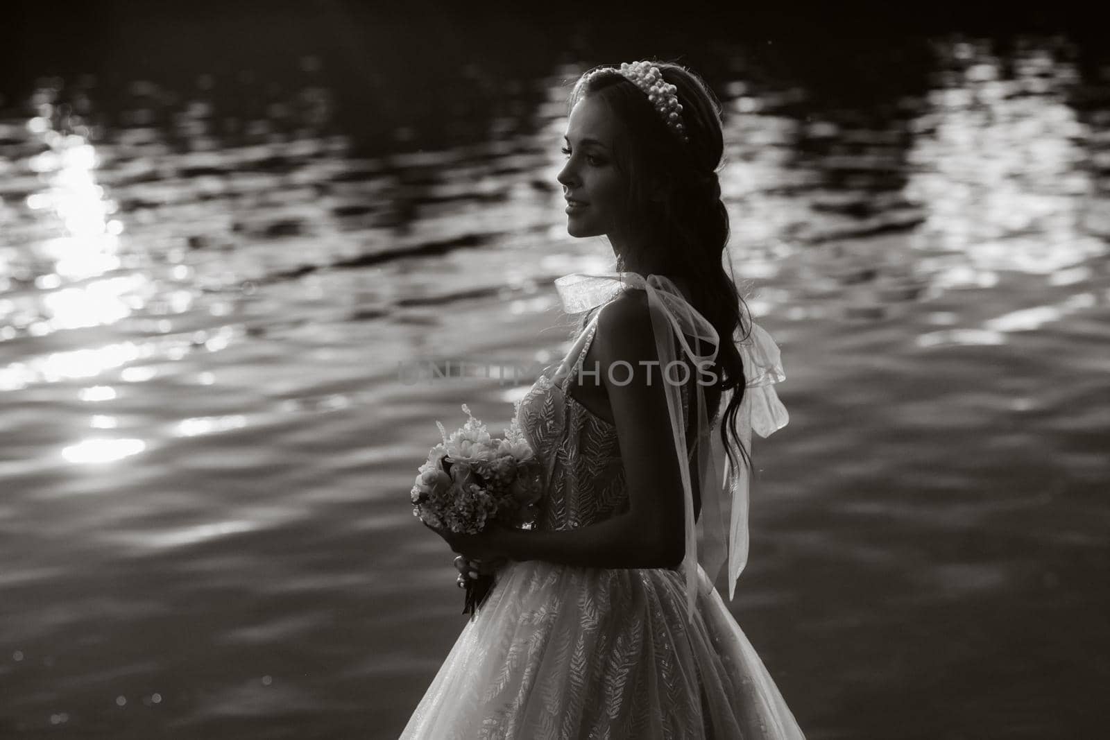 An elegant bride in a white dress and gloves stands by the river in the Park with a bouquet, enjoying nature at sunset.A model in a wedding dress and gloves in a nature Park.Belarus. black and white photo. by Lobachad