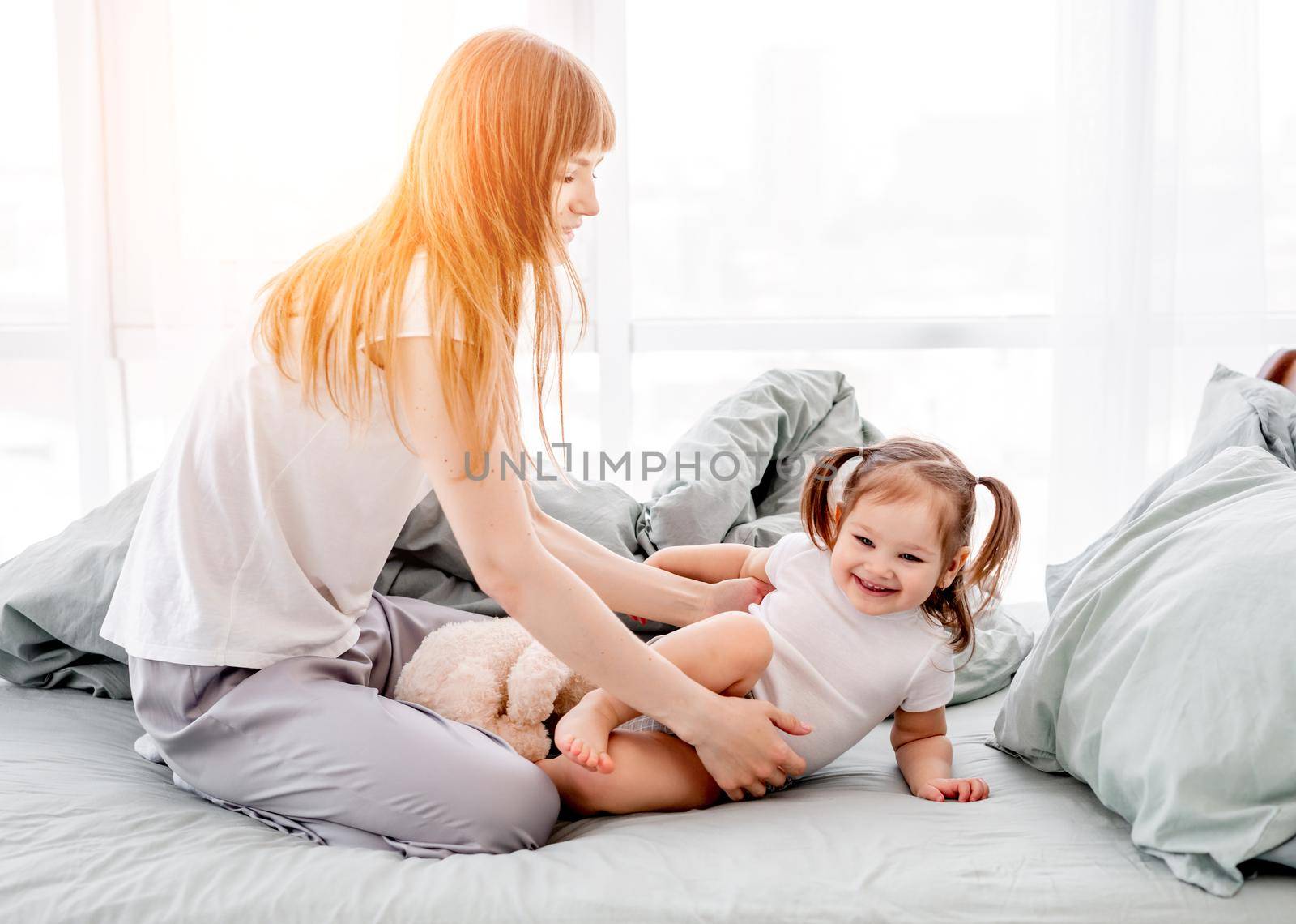 Beautiful mother playing with little daughter in the bed in the room with sunlight. Happy family moments of mom with her child. Attractive girl with her kid in the bedroom