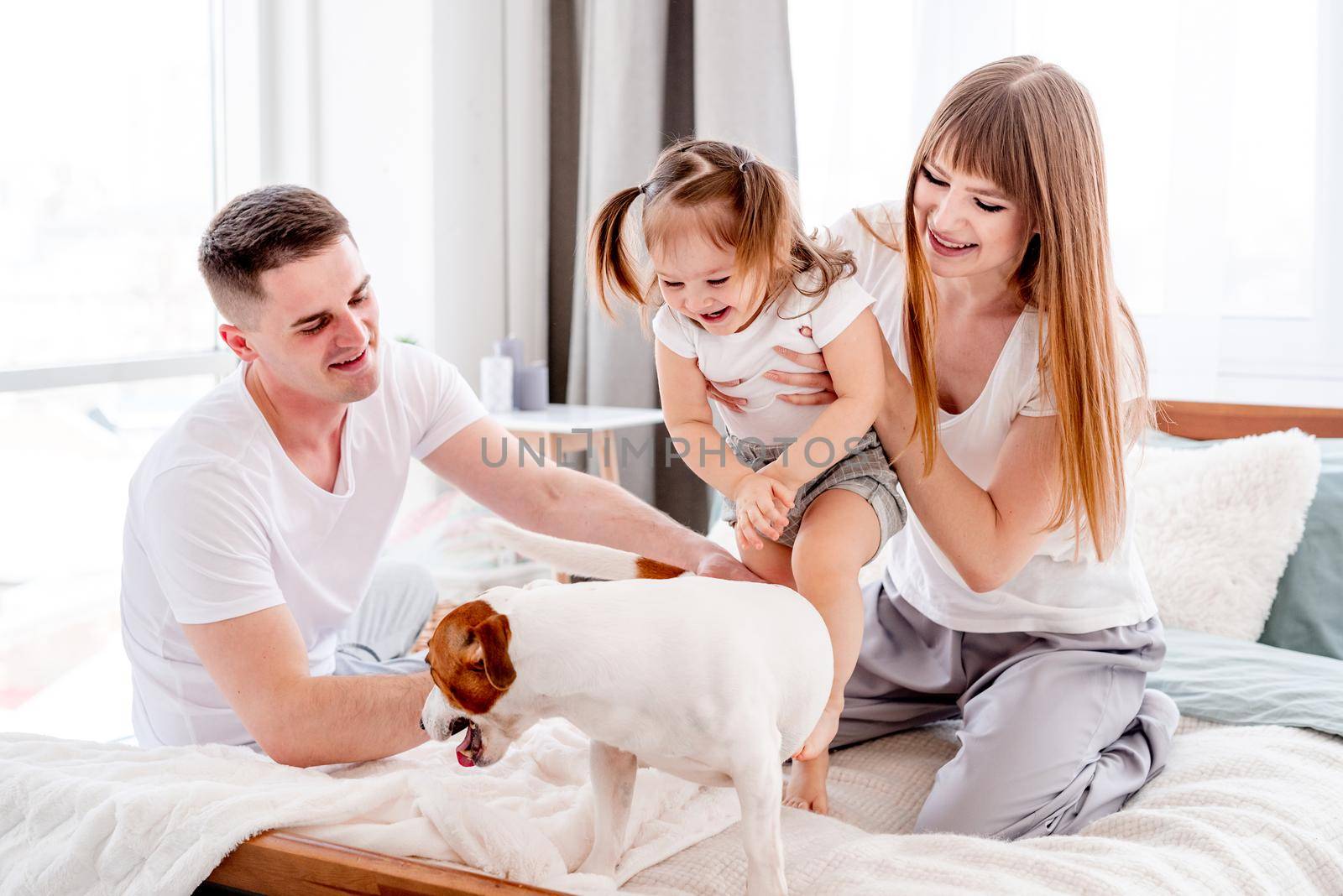 Sunny family morning in the bedroom. Beautiful parents with their daughter staying in the bed with dog. Mother, father and child wearing pajamas in the morning with pet