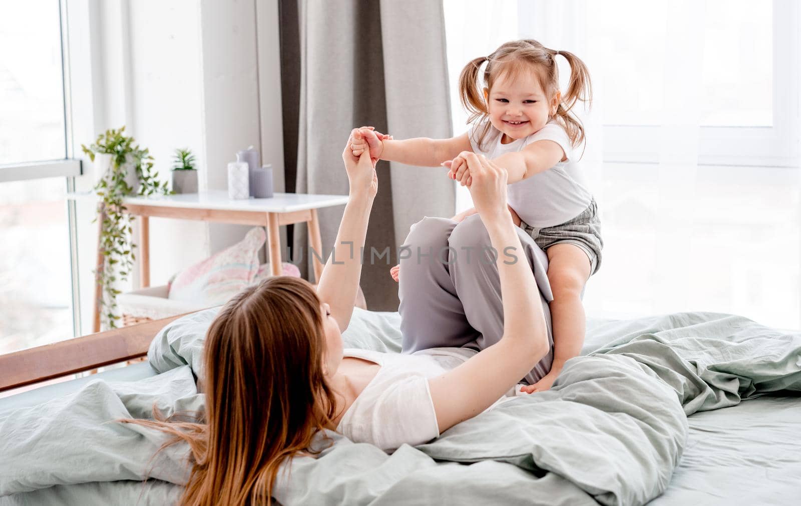 Young mother playing with daughter in the bed swinging cute child on her legs. Caucasian mom and her kid spend time together in the bedroom with sunlight. Beautiful family time