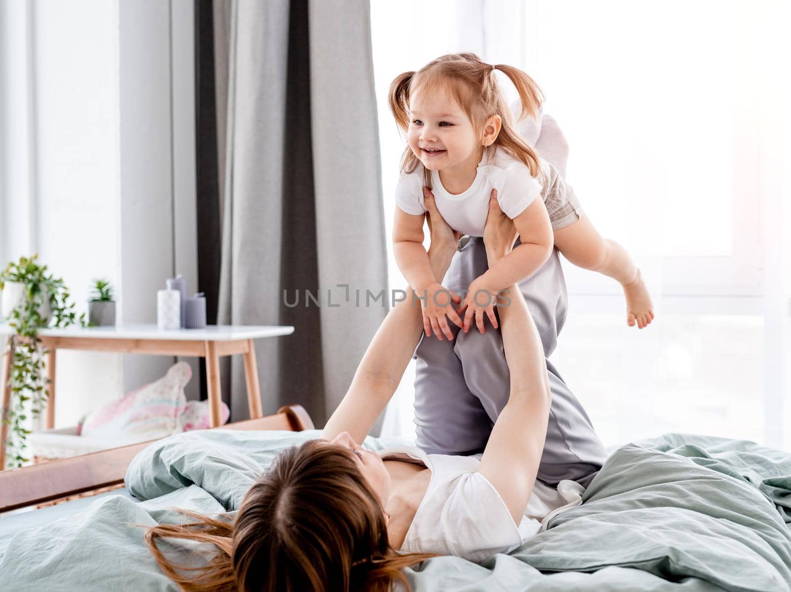 Young mother playing with daughter in the bed holding child on her legs. Mom and daughter spend time together in the bedroom with sunlight. Beautiful family time