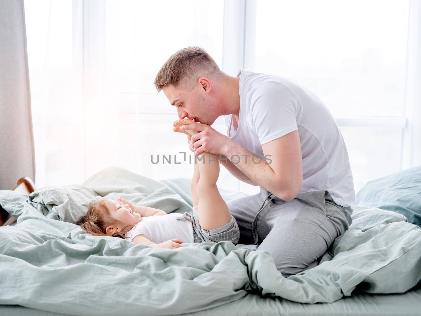 Young father kisses feet of her little daughter lying in the bed. Smiling child with her parent in the morning time. Beautiful moments of dad with his kid in the bedroom together