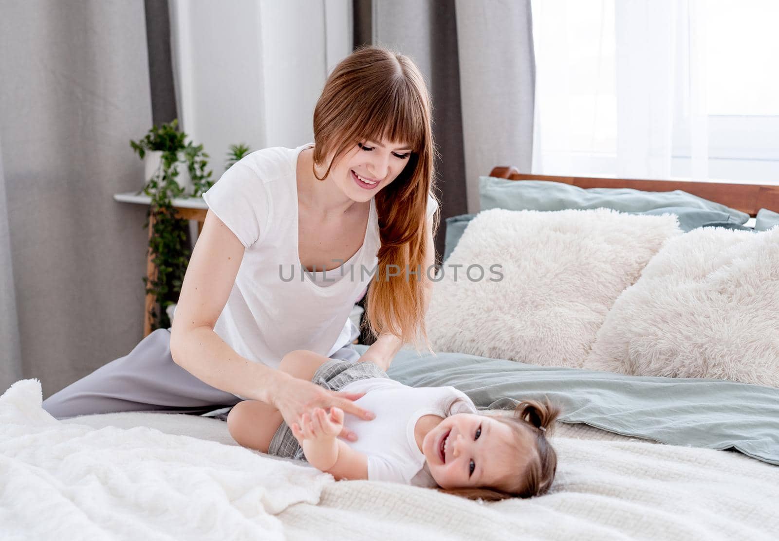 Beautiful young mother sitting in the bed wearing pajama with little daughter and smiling. Attractive girl mom playing with her child in the bedroom. Morning family moments
