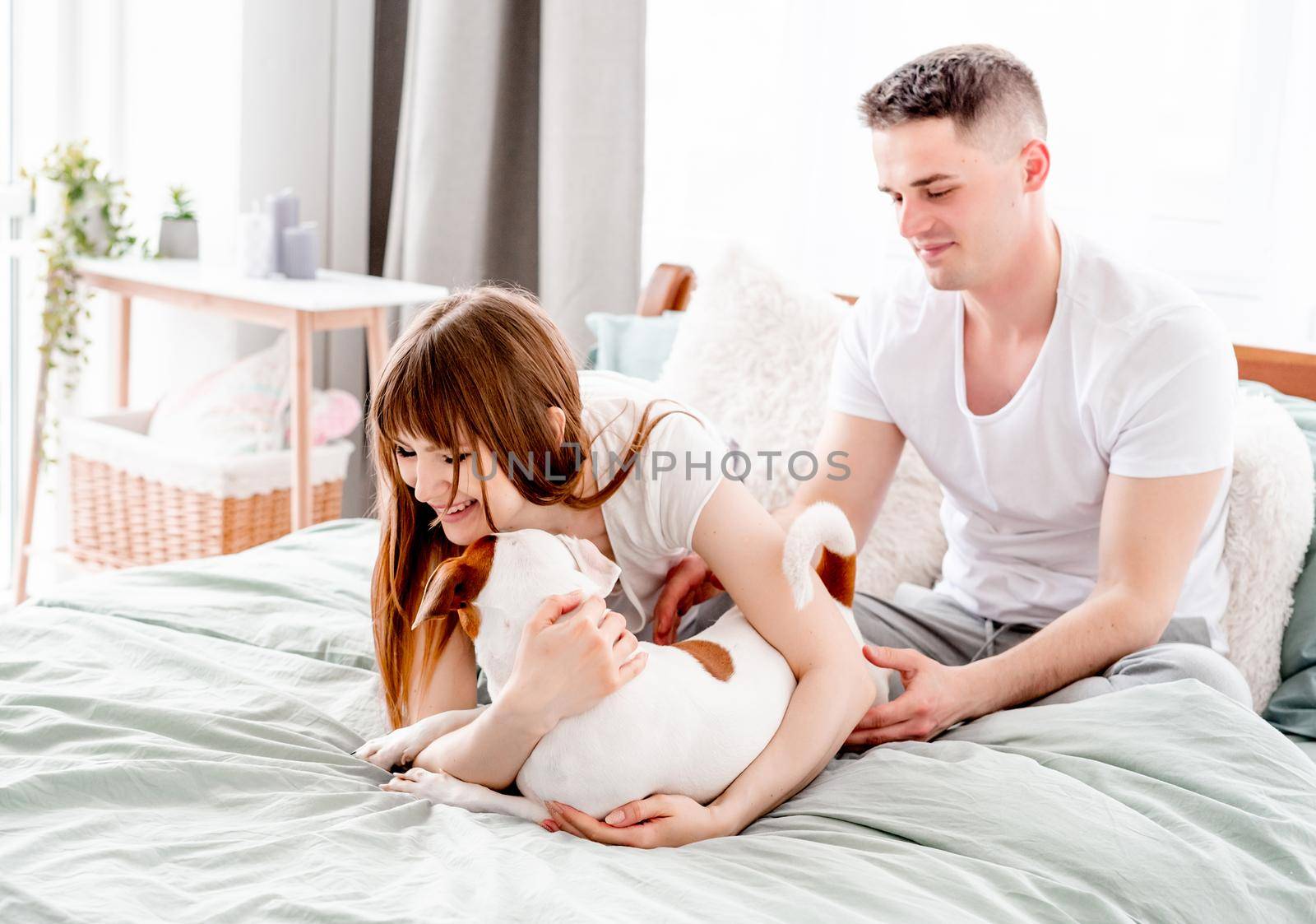 Beautiful couple spend time in the bed together and petting their cute dog. Family morning moments. Attractive girl hugging pet and smiling. Young wife and husband with their doggy