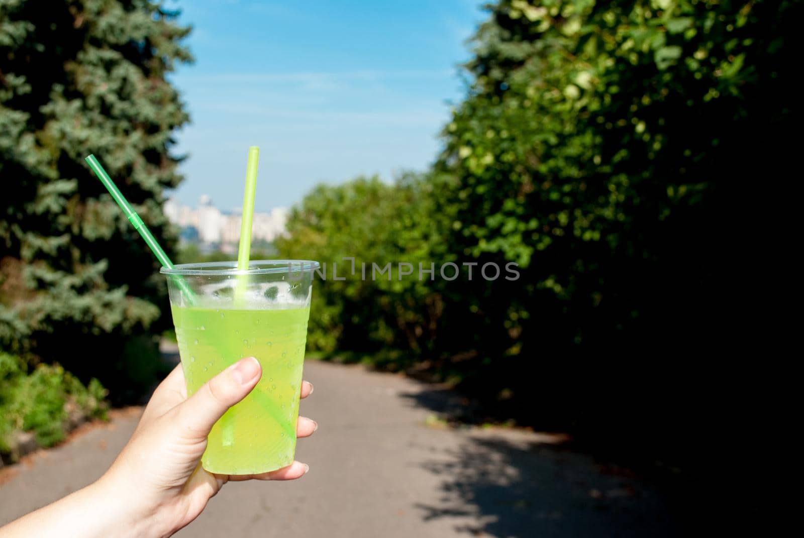 Woman holding plastic green cocktail cup close-up, outdoors in summer in the park.