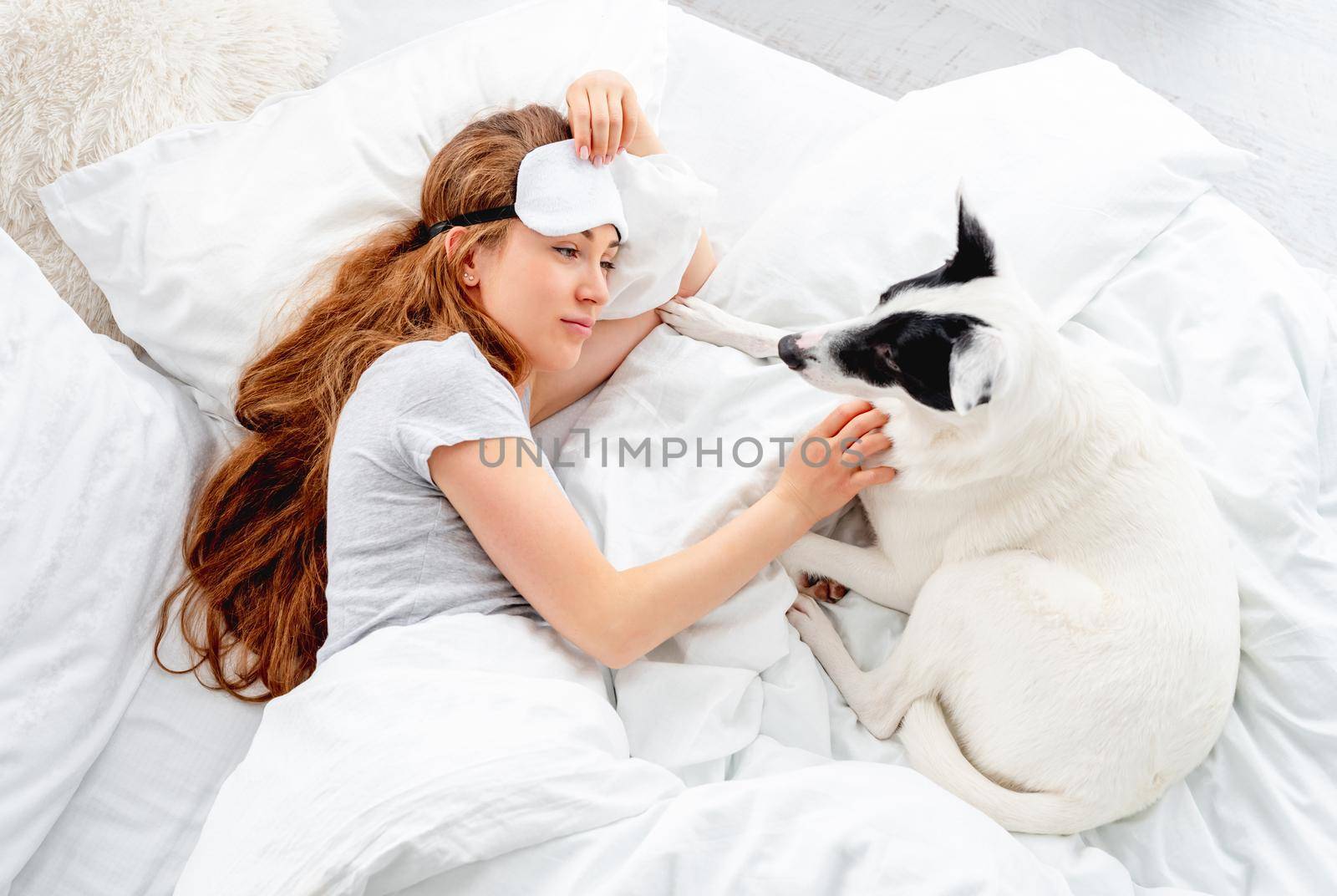 Beautiful long hair girl lying in the bed with cute dog and petting him. Pretty young woman resting with pet. Female person wearing eye sleeping mask with doggy