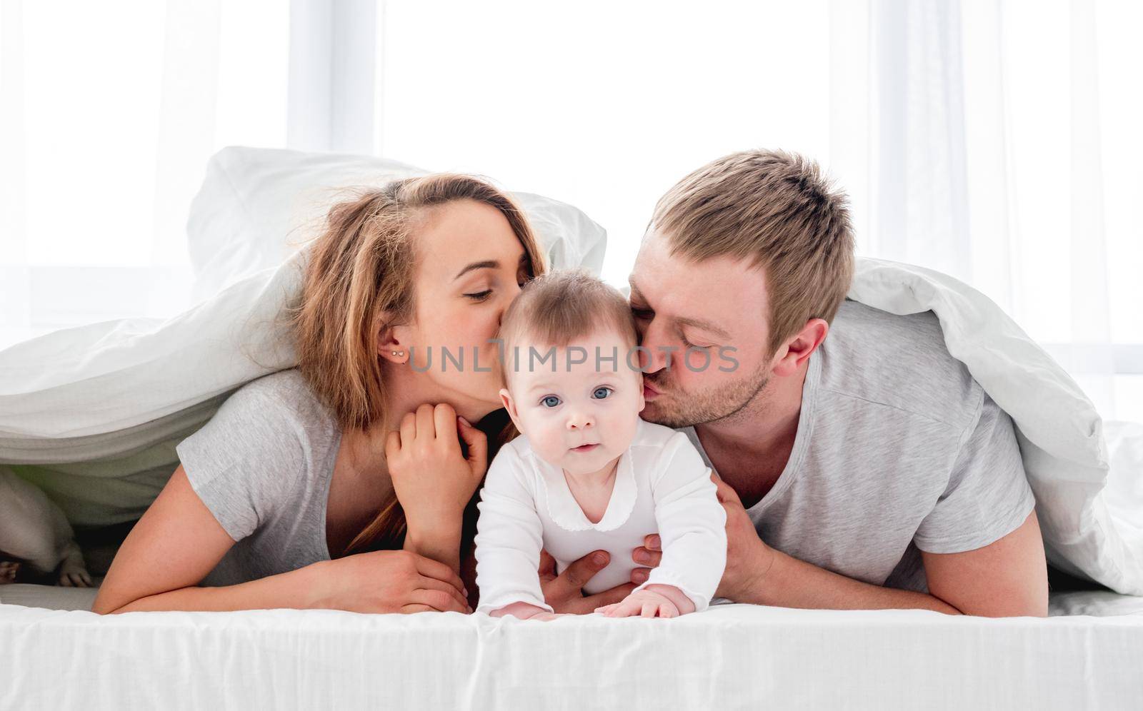 Mother and father lying in the bed under blanket with their son and kissing baby on his cheeks. Beautiful family morning together in sunny bedroom. Happy parenthood