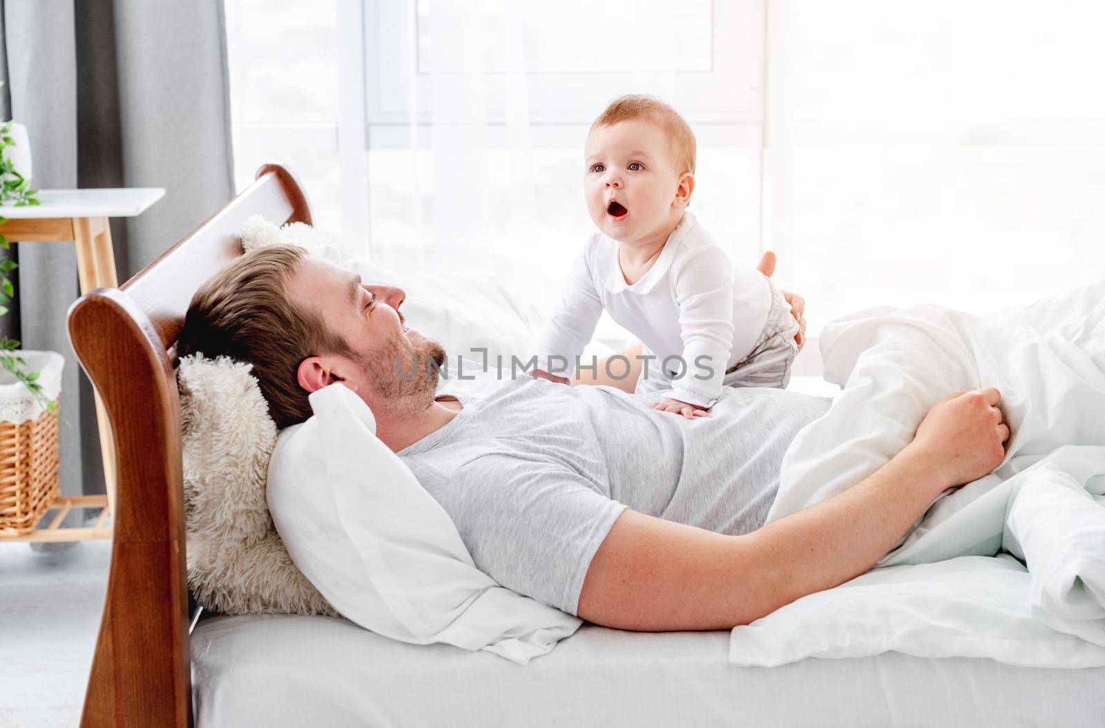 Young father lying in the bed with little son in the sunny morning time. Man with his child baby boy resting in the bedroom. Happy parenthood