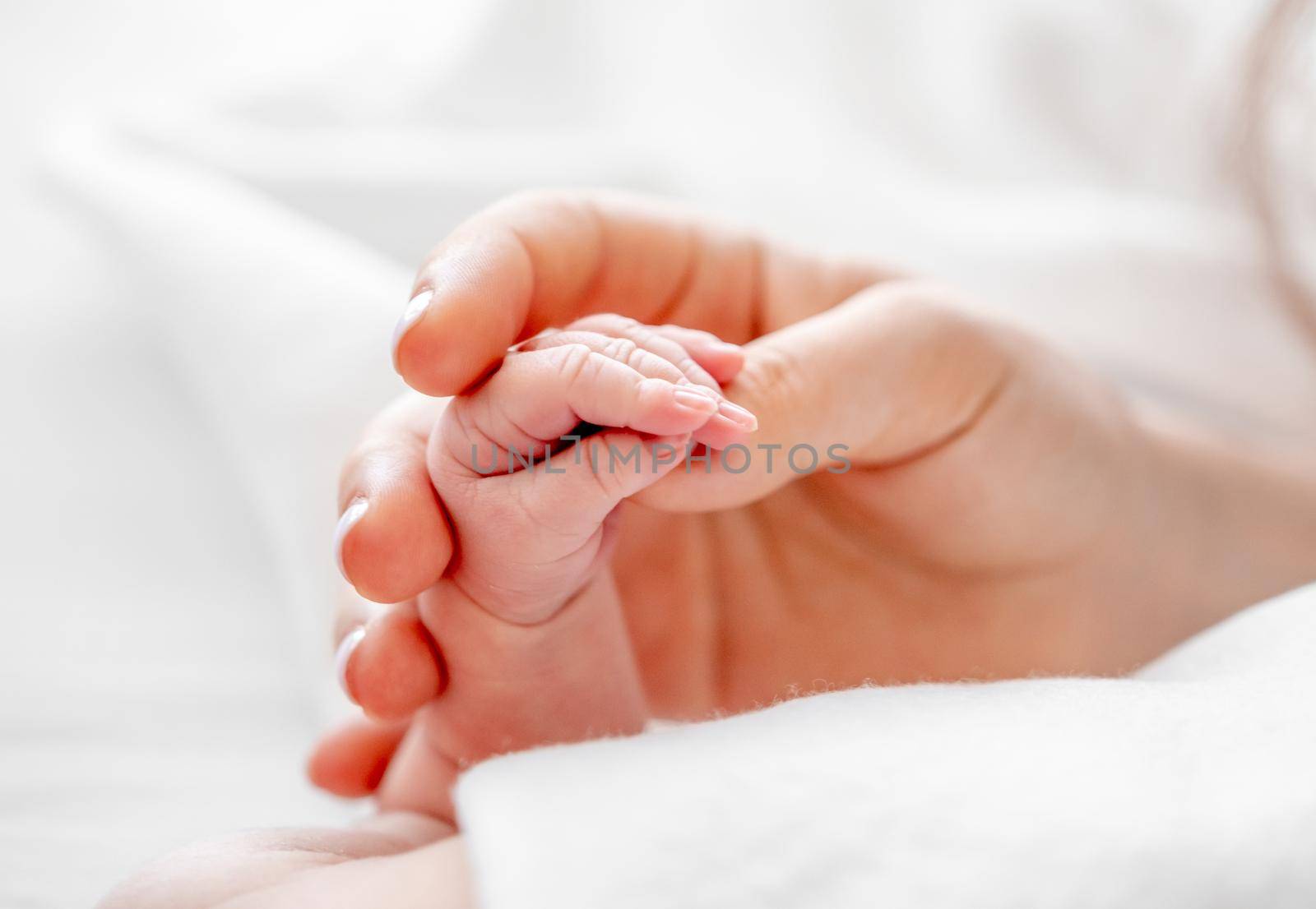 Mother holding tiny newborn baby hand with little fingers closeup in the bed with white bedding with daylight. Concept of matherinity love, protection and tenderness