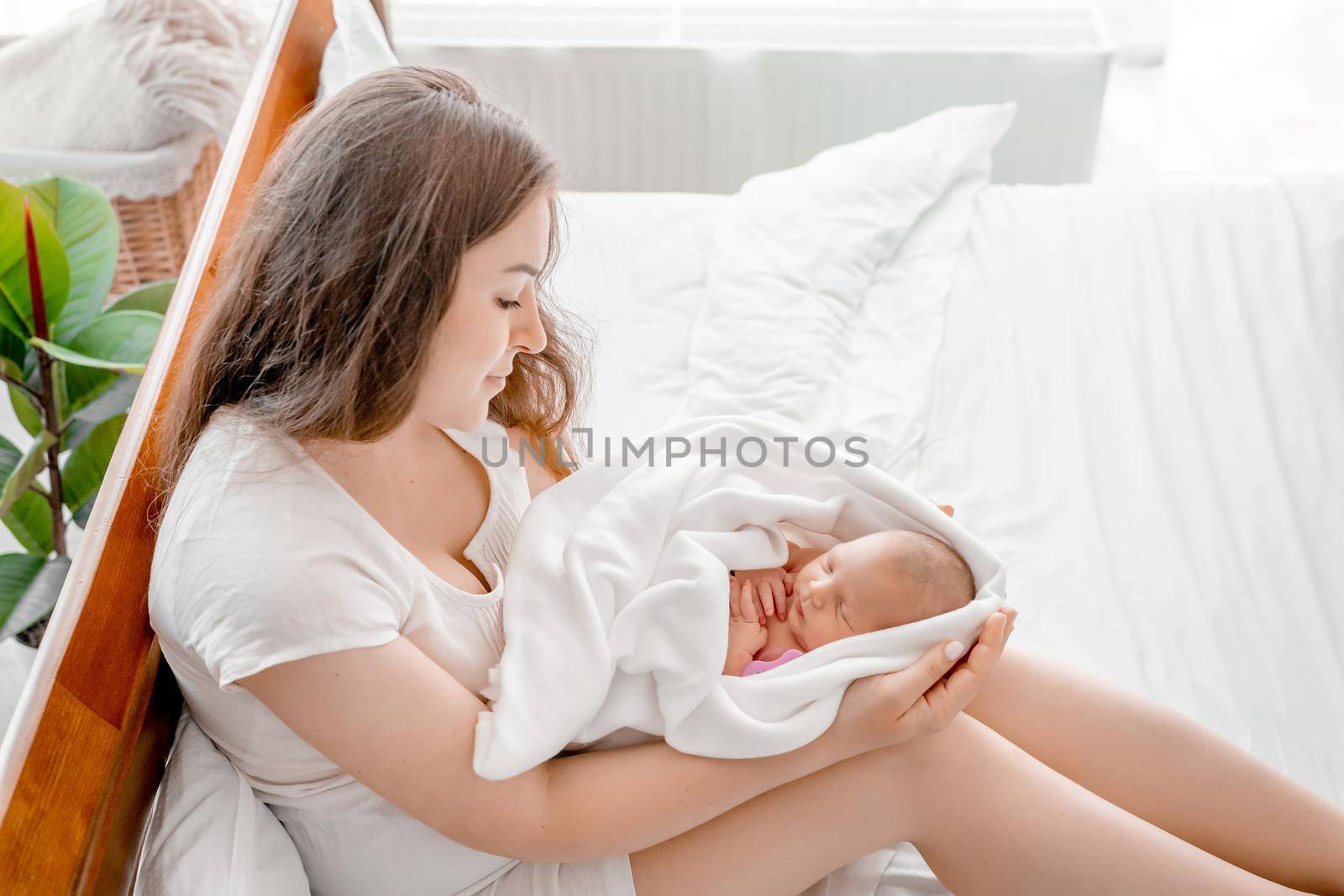 Beautiful young mother with newborn daughter in the bed with daylight together. Adorable infant baby girl sleeping in hands of her mother