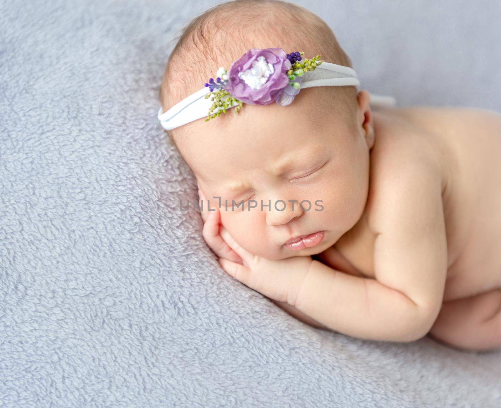 Naked baby girl wearing flowery hairband sleeping on her side, hands pressed to the cheek