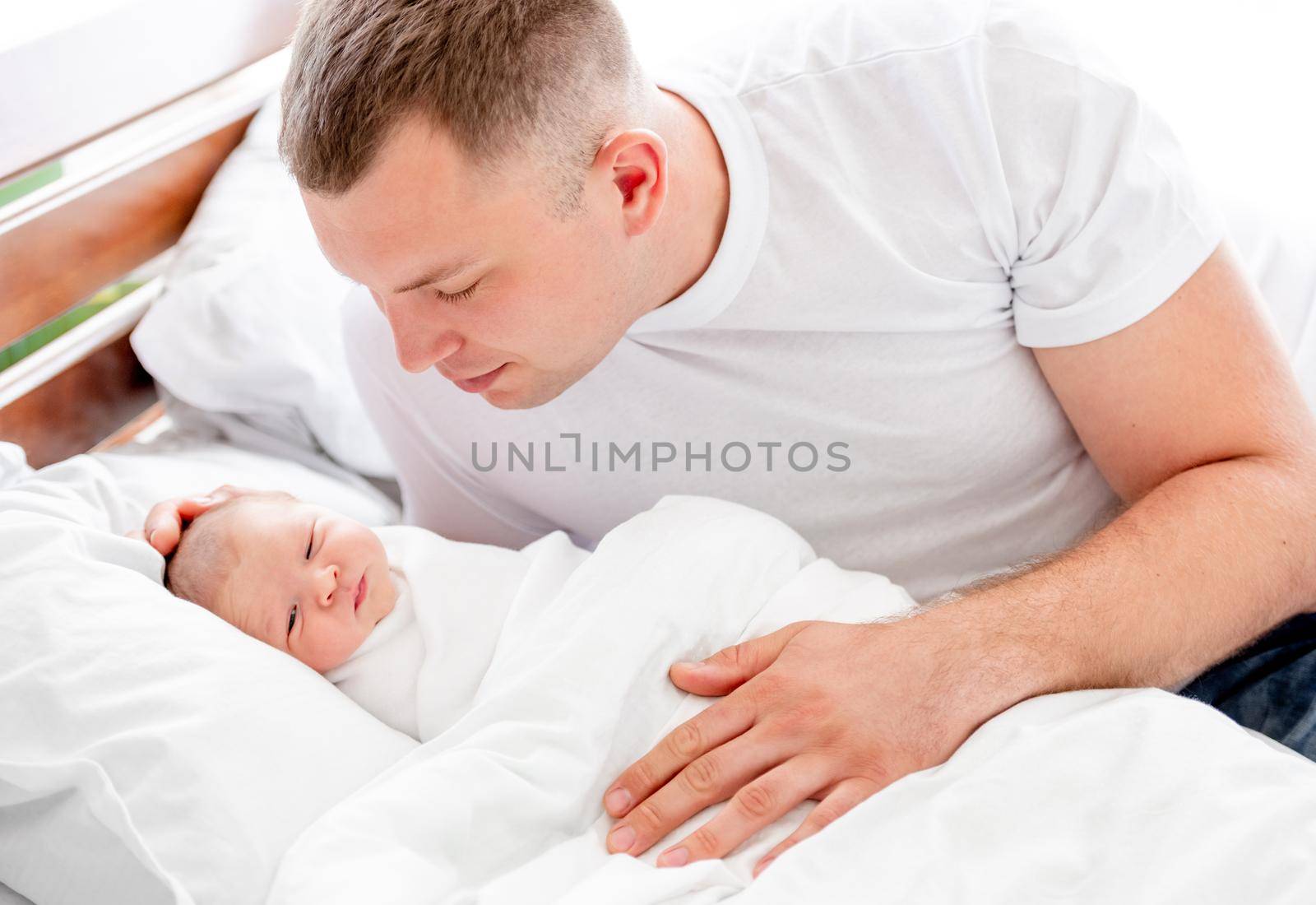 Young father sitting in the bed and looking at his newborn baby daughter sleeping in white sheets. Parent dad and napping infant child at home with daylight