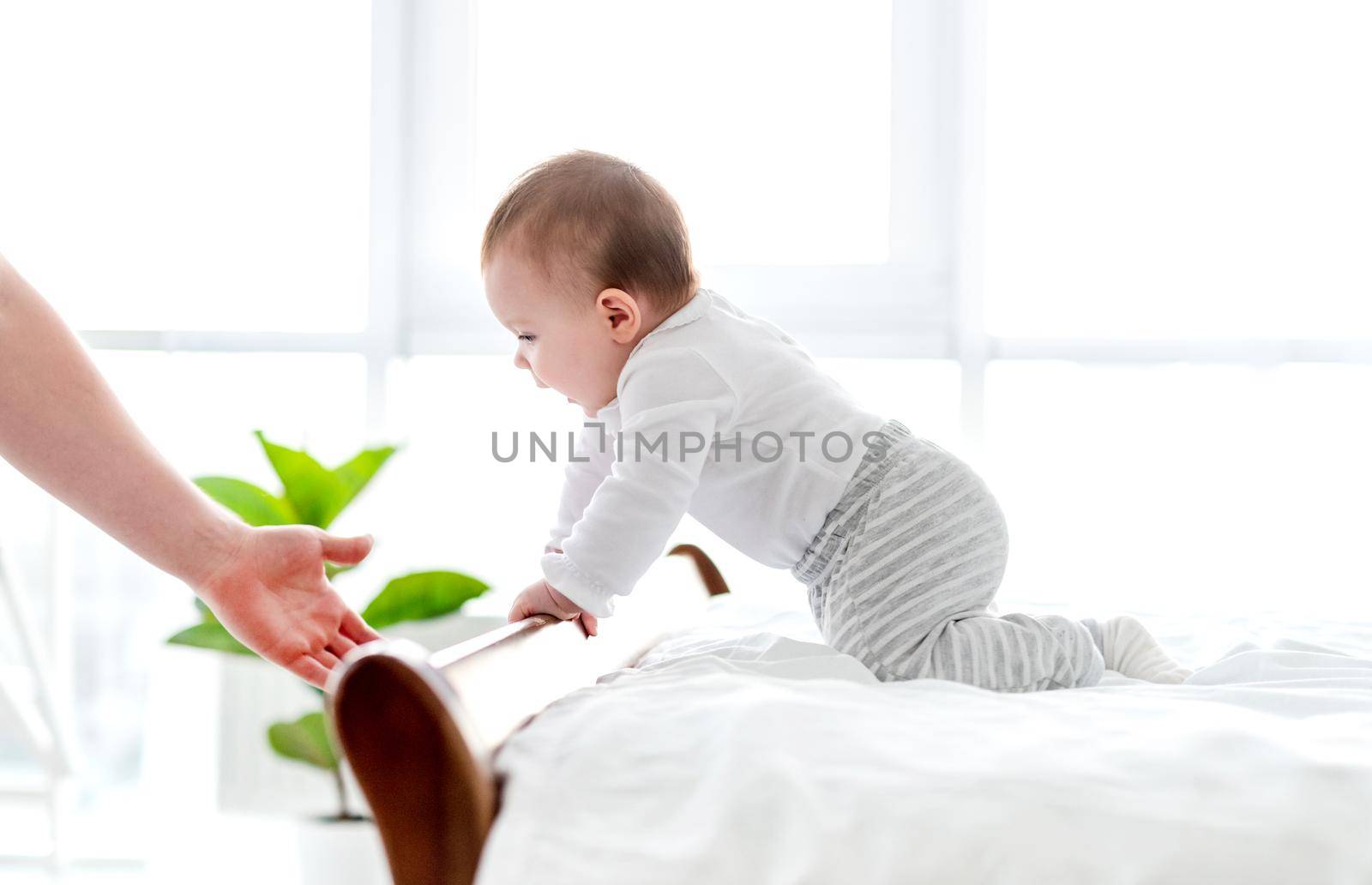 Adorable little baby boy creeps in the bed in the room with panoramic windows and parent hand stops him preventing falling. Beautiful toddler child in the bedroom in the morning time. Cute kid at home