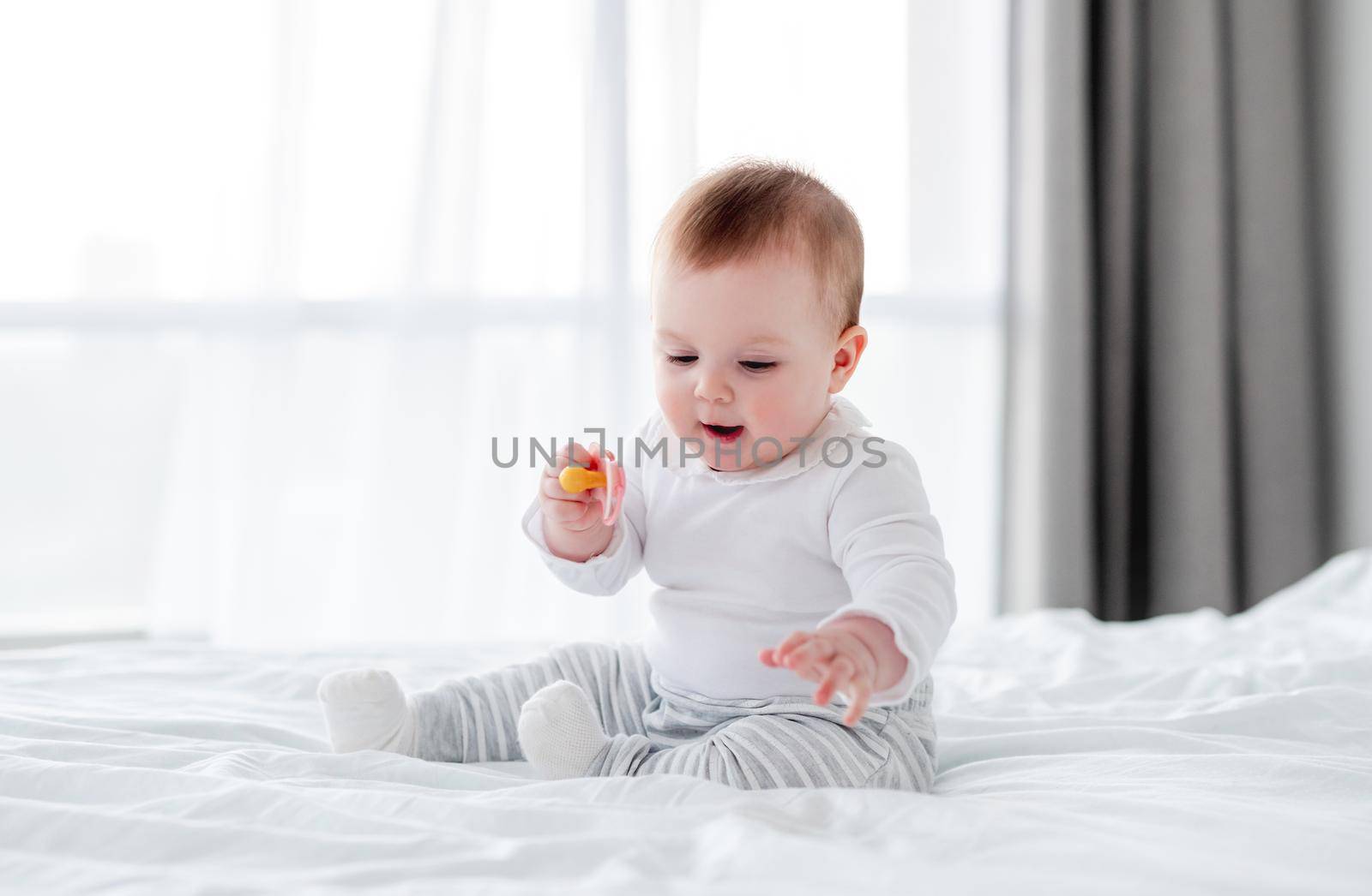 Adorable little baby boy sitting in the bed and holding dummy in his hand. Beautiful toddler child in the bedroom in the morning time. Cute kid alone at home