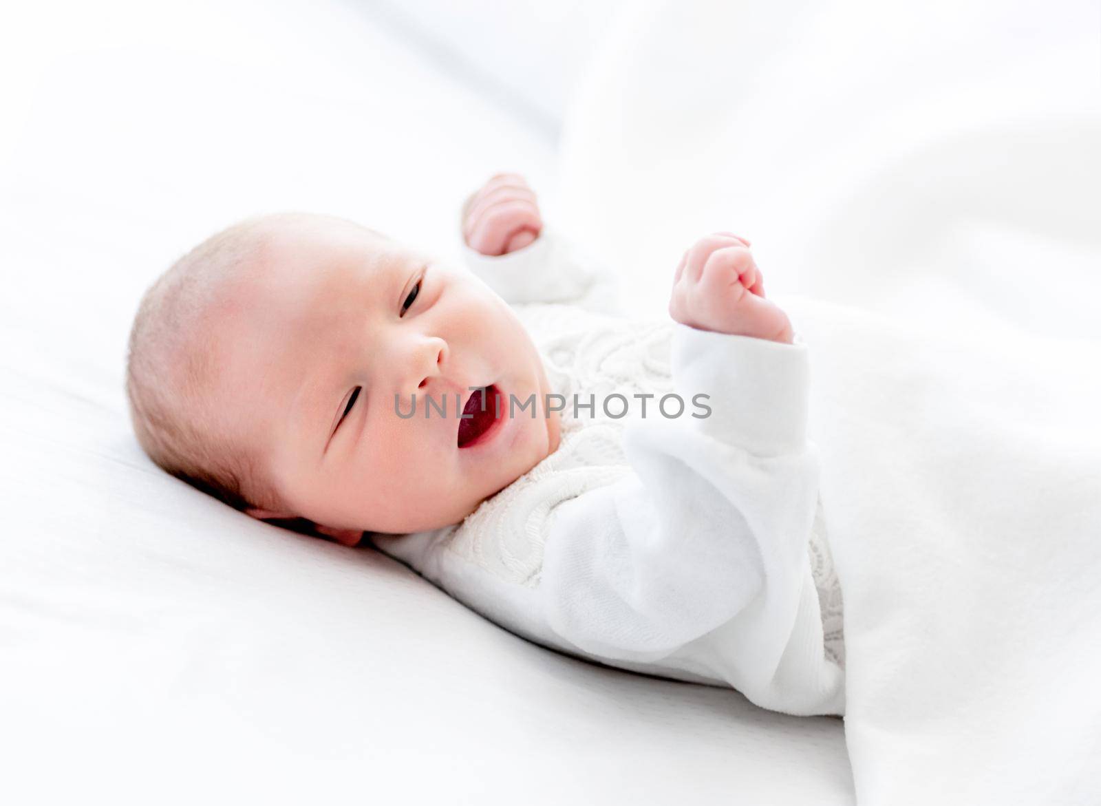 Closeup portrait of adorable newborn baby girl wearing white costume lying in the bed with mouth open. Cute infant child resting at home