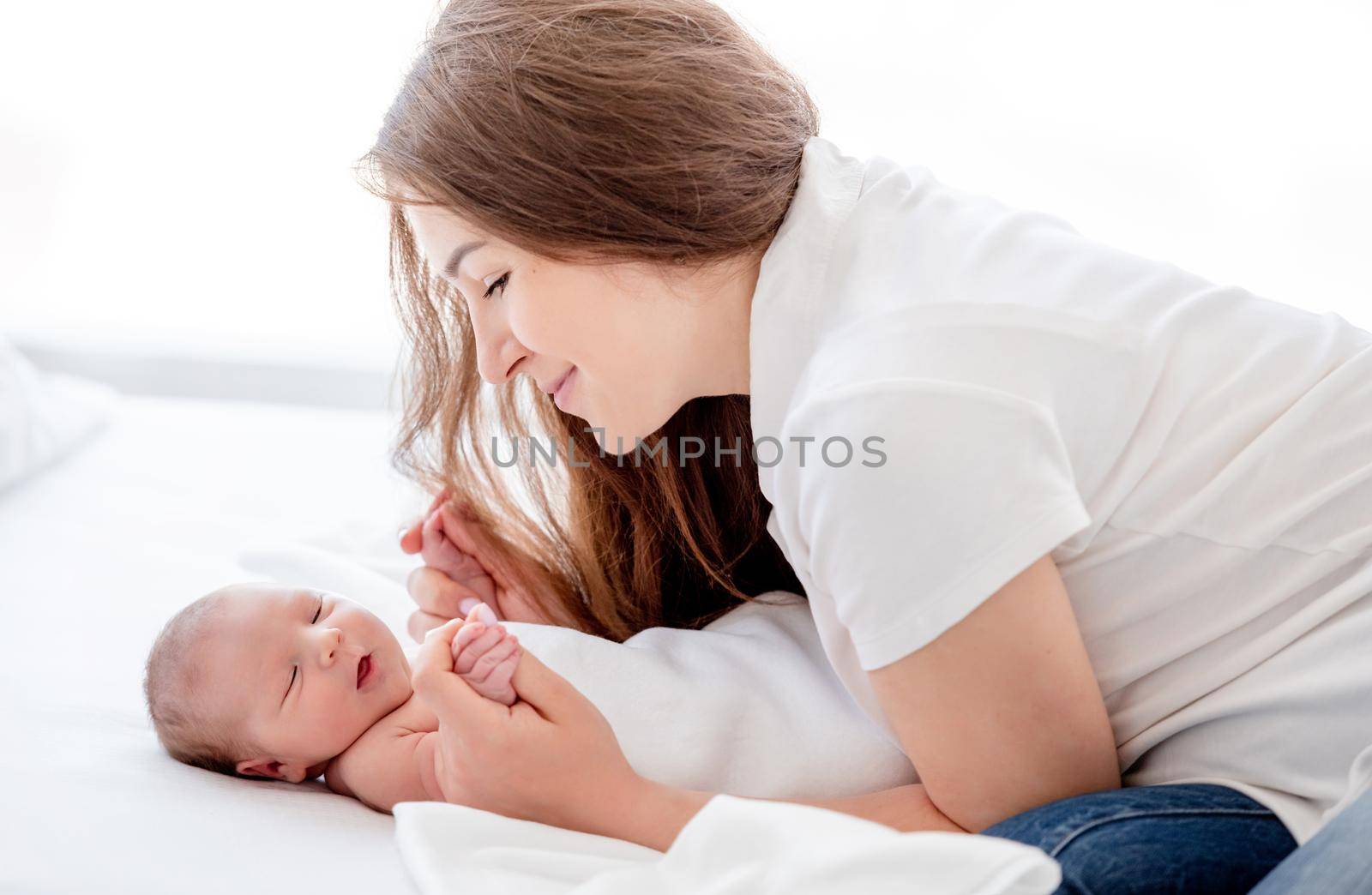 Beautiful young mother holding hands of her sleeping newborn baby daughter in the bed and looking at her with smile. Parent and infant child. Family moments of matherinity