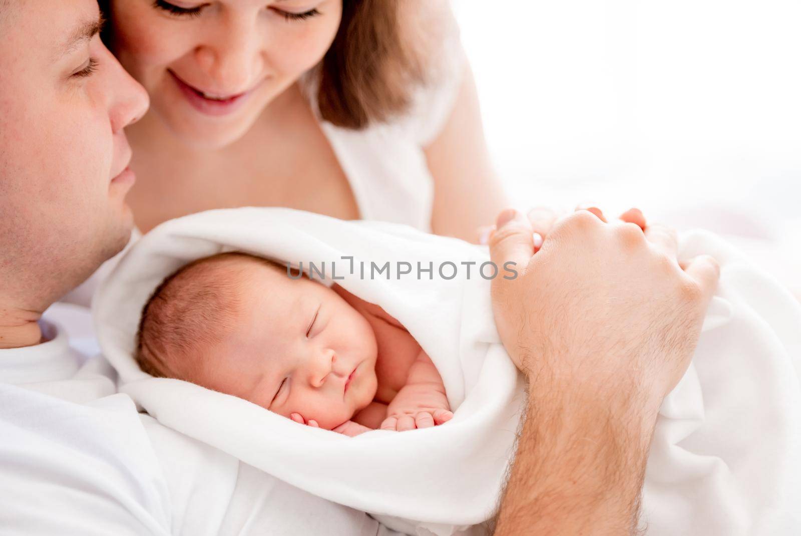 Young mother and father holding in hands sleeping newborn baby girl. Parents with infant daughter in the bed. Warm family moments