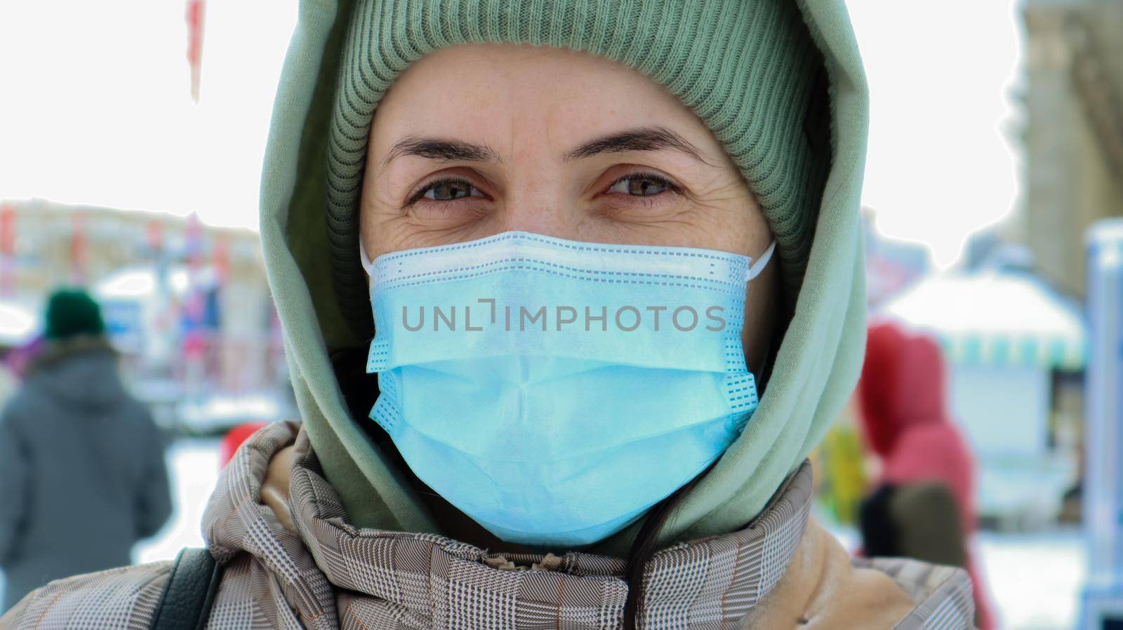 Portrait of happy young woman wearing medical protective mask outdoors and looking at camera. Virus protection. A woman outdoors on a winter day stands in a hat and a hood in the park