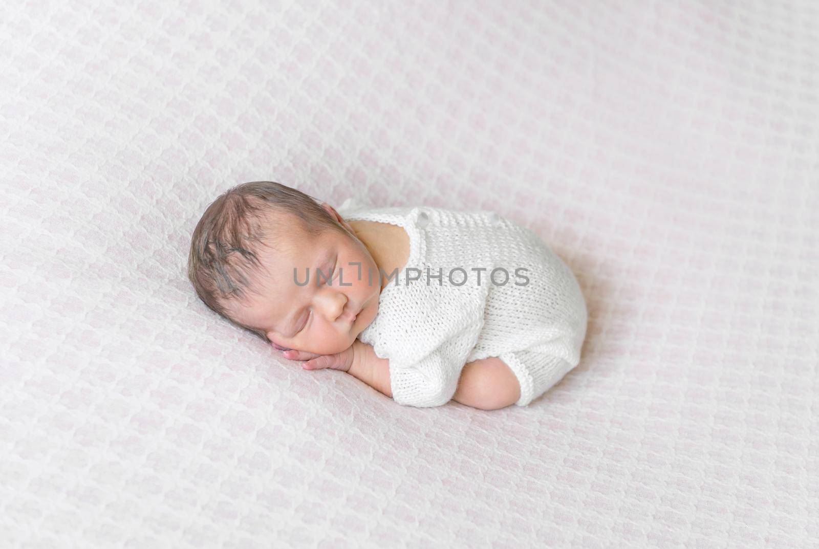 Sweet baby sleeping curled up on his belly, wearing white suit, on pink soft surface