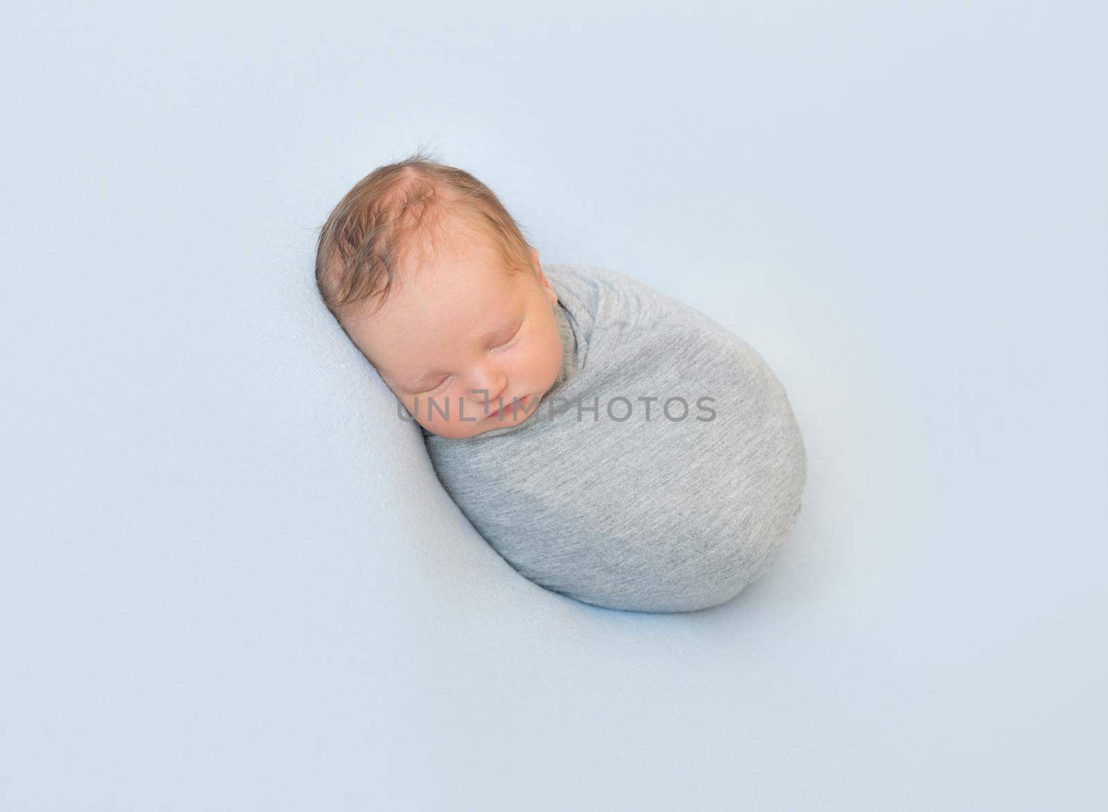 Tiny hairy infant sleeping curled up, wrapped in gray small blanket, on side, blue background