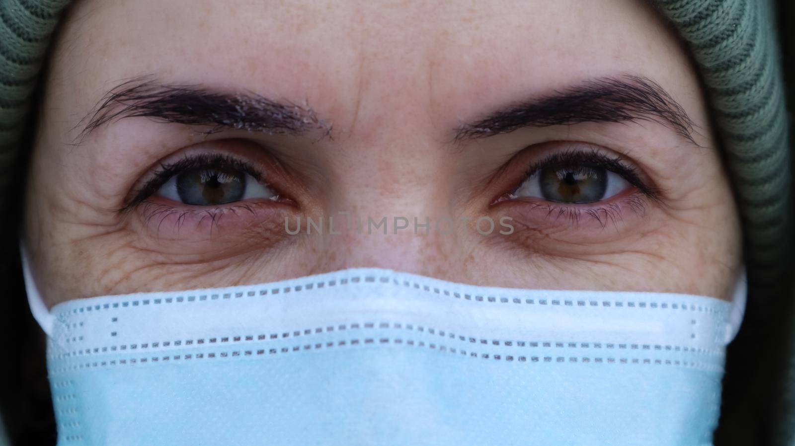 A beautiful young woman with a medical mask on her face is looking at you in the camera. Close-up portrait, fear in the eyes. Perfect eyes. Winter. Coronavirus protection. Health and safety concept