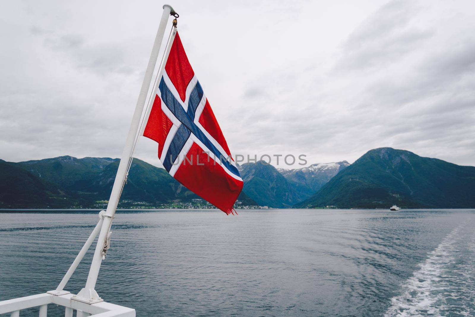 Norwegian flag hanging on the railing of the ship and waving above the water. Norvegian fjord with a flag. Ferry trip in Norway. Norway Flag on sea and mountains background.