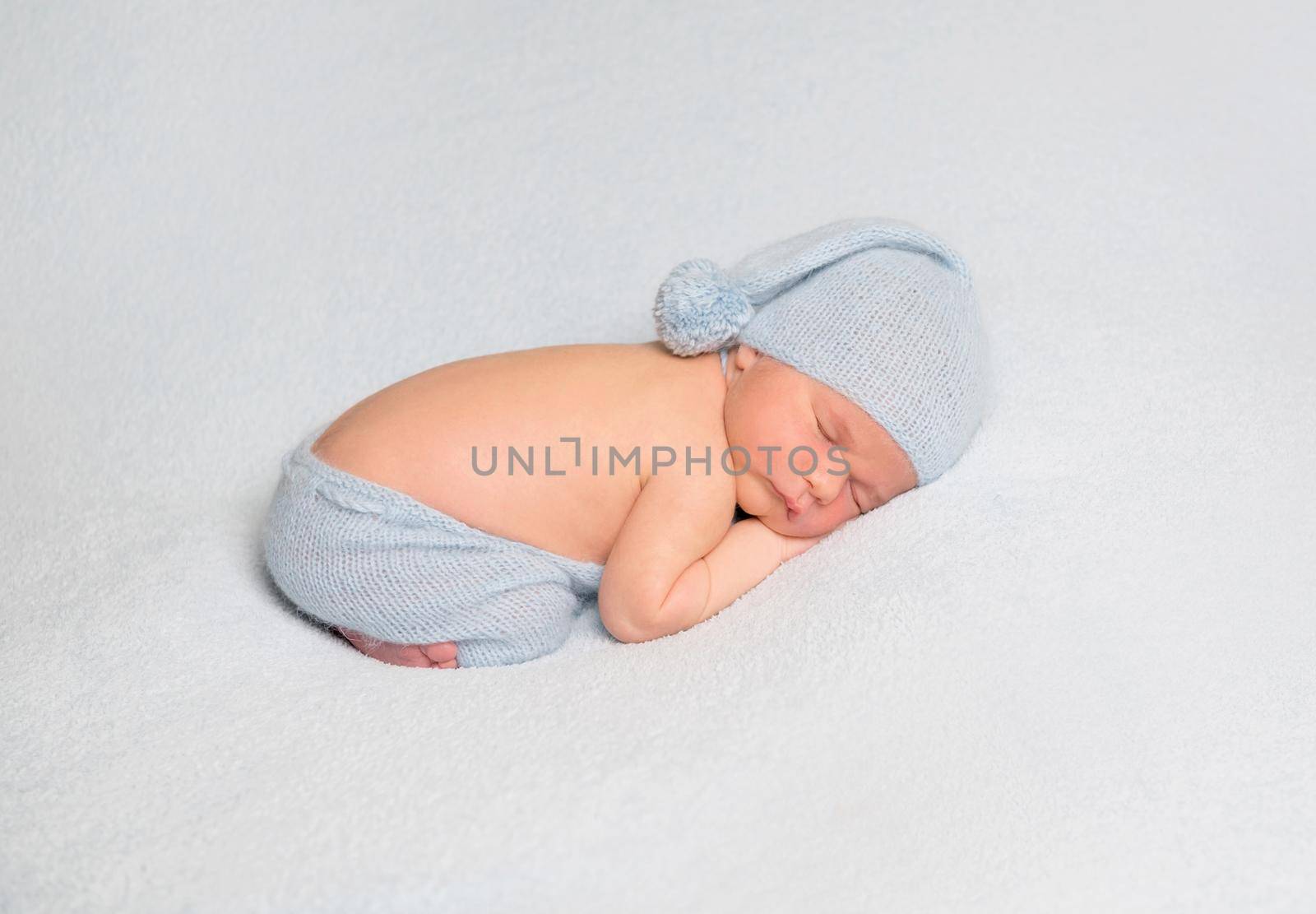 Sleeping baby boy in blue bonnet curled up on white blanket.