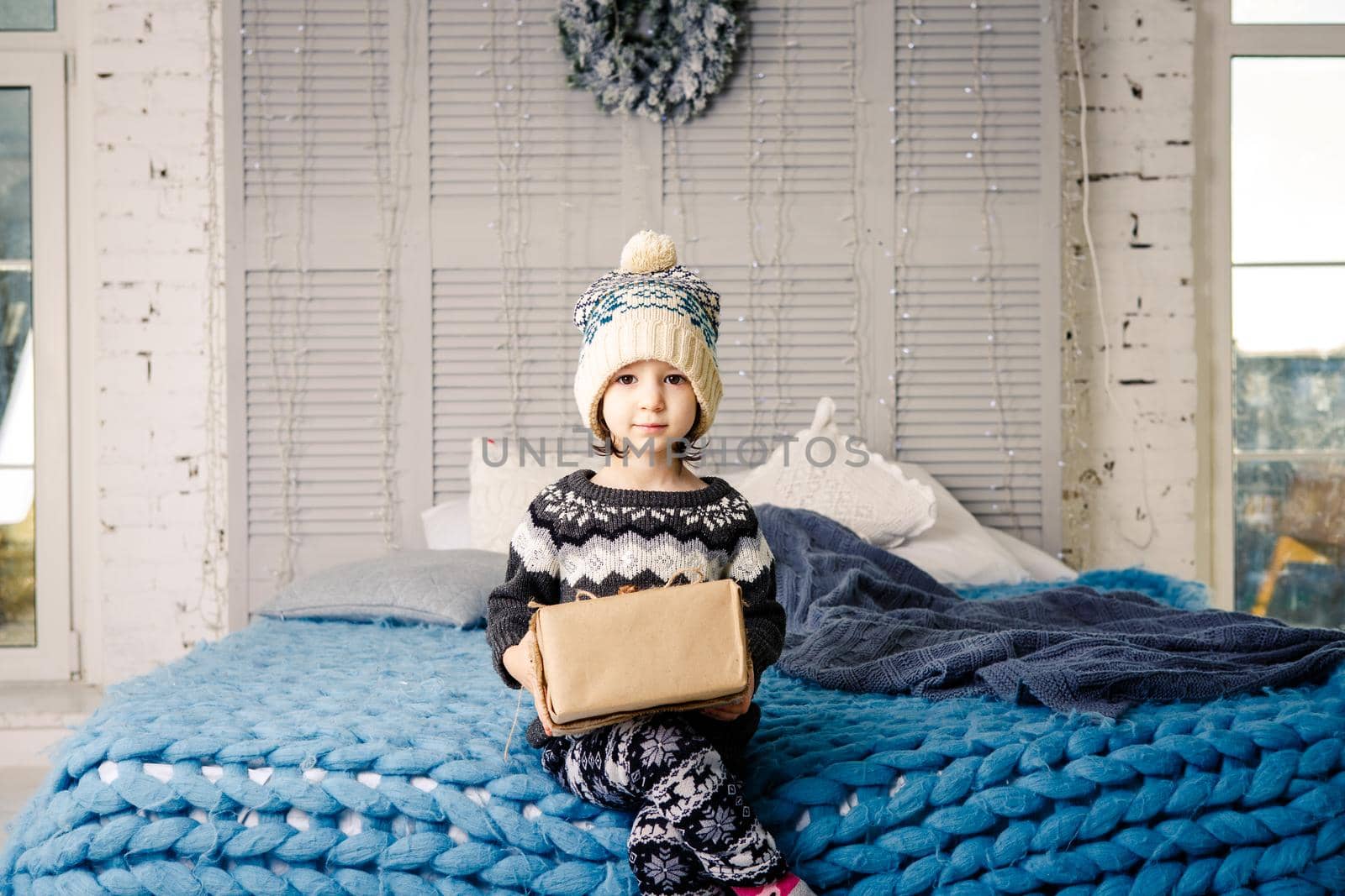 little girl the child sitting in pajamas and hat on the bed with garland of light bulbs with gifts boxes wrapped in a non-colored paper decorated with cones on blue knitted coverlet.Christmas concept by Tomashevska