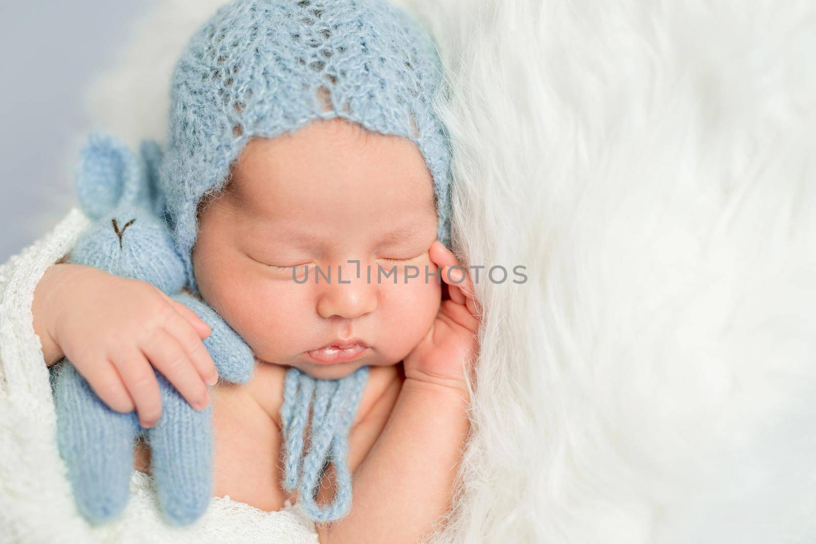 sleepy newborn boy in blue knitted hat with toy on white fluffy blanket