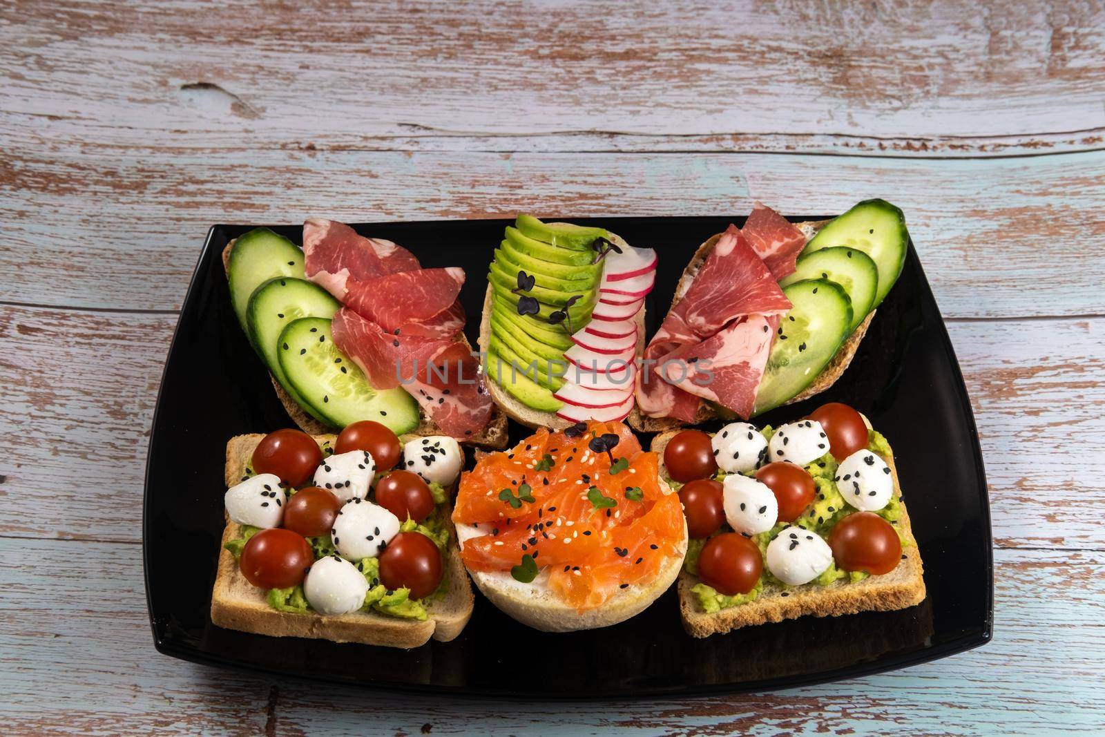 Assorted sandwiches with fish, cheese, meat and vegetables on a black plate and wooden background.