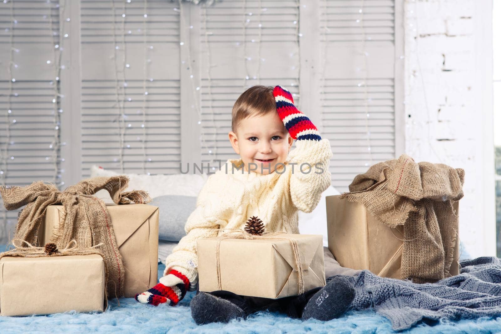 little funny playful boy a child sits on a bed on Christmas day with gift boxes in white wool knitted sweater and big bright mittens on it and laughs out loud. In interior there is a festive decor.