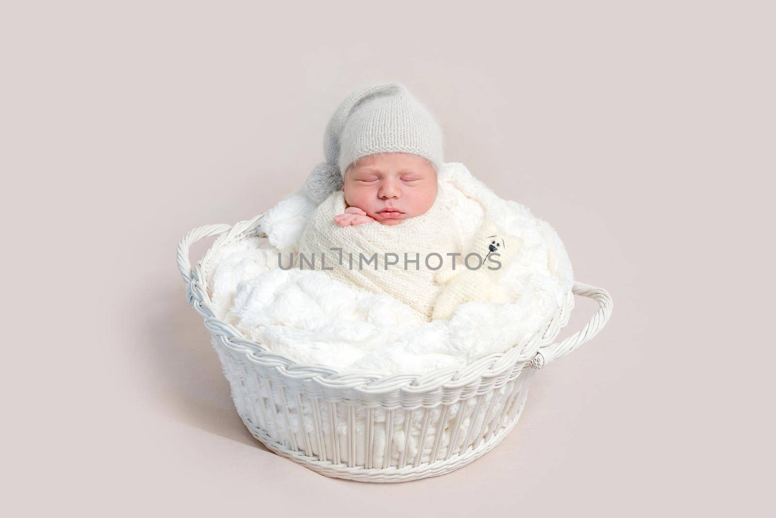 Top view of newborn baby wrapped in white wrap wearing white bonnet and lying on white round basket with soft blanket. Newborn baby in white outfit