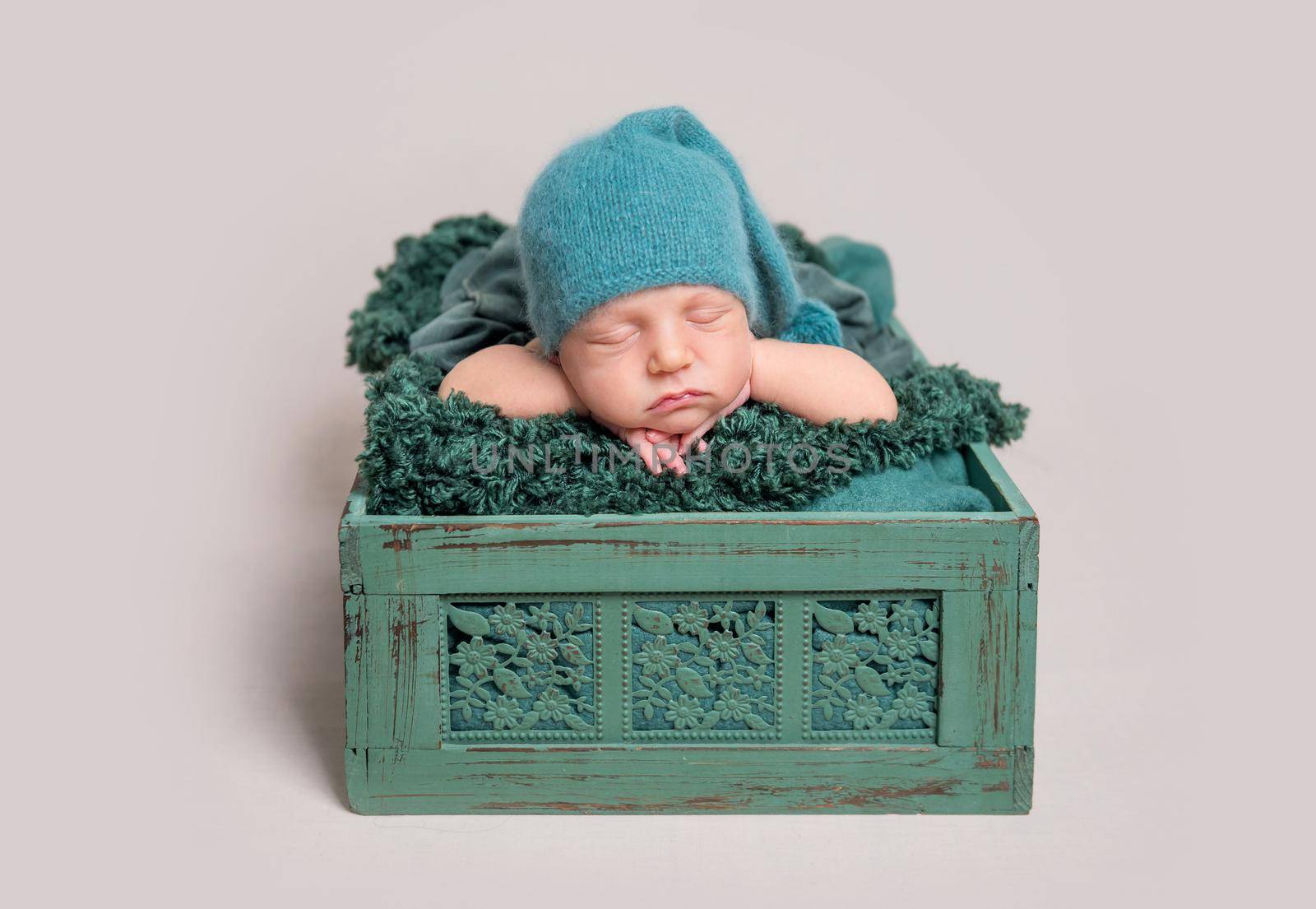 Beautiful newborn baby in green cap lying in wooden crate with arms under the head