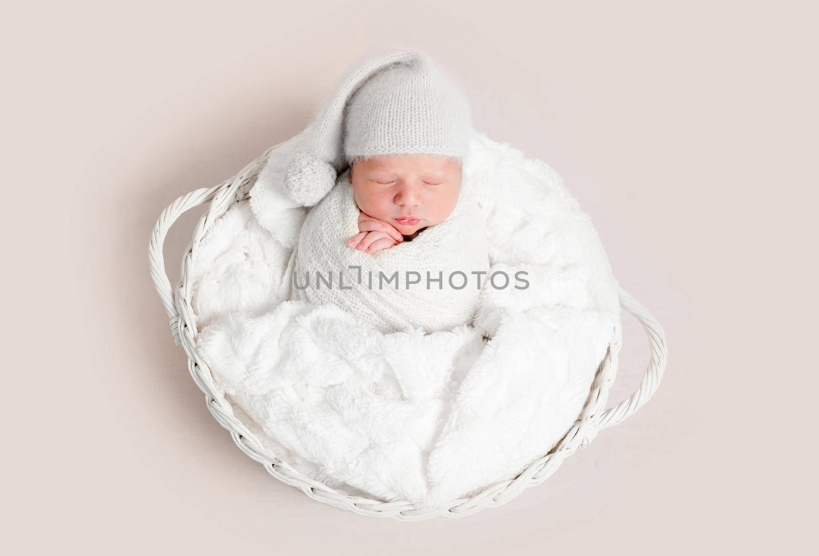 Top view of newborn baby wrapped in white wrap wearing white bonnet and lying on white round basket with soft blanket. Newborn baby in white outfit