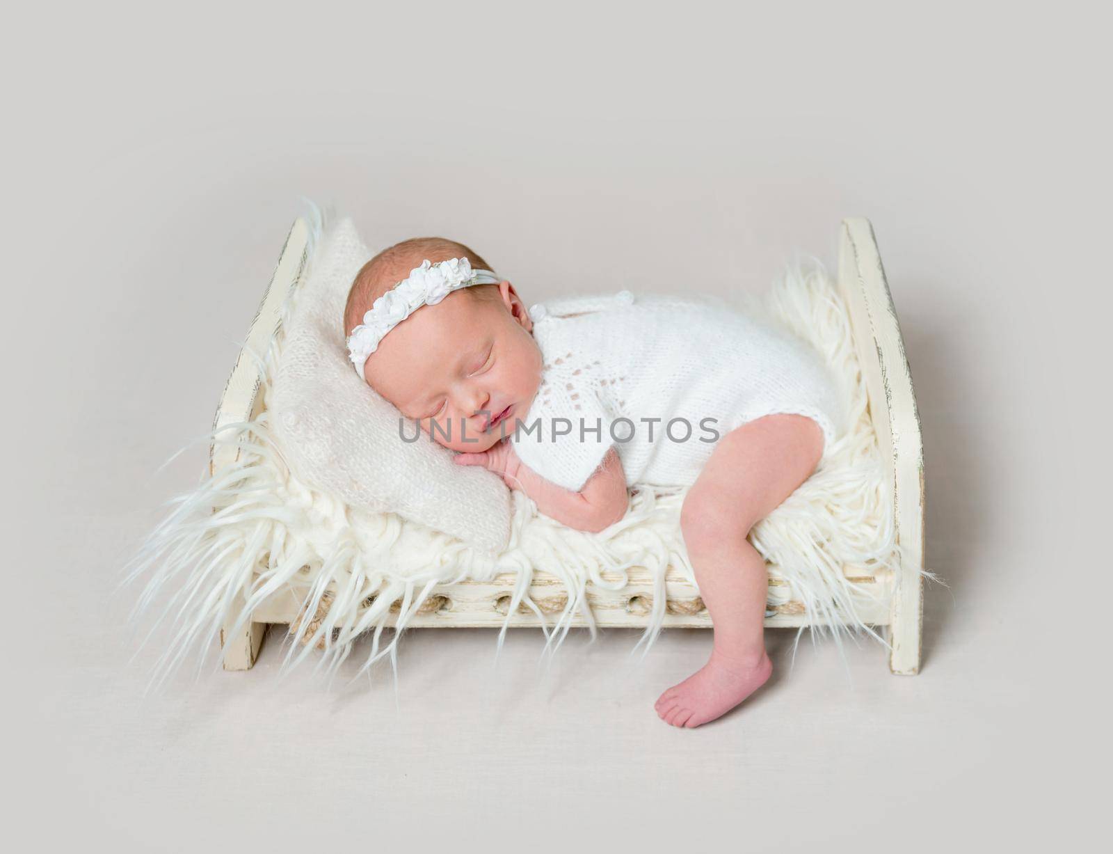 Lovely newborn baby girl sleeping on small crib lowered her small leg on the floor. Little child girl in white outfit with flowery headband