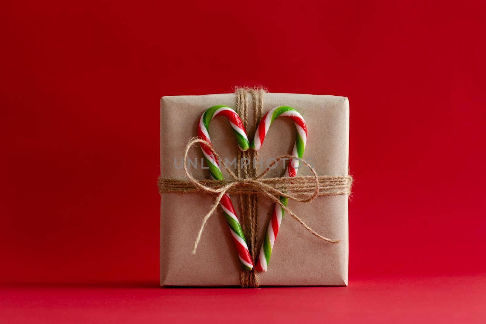 Gift box packed in kraft paper on a red background. Heart of Christmas lollipops in the form of a cane. Bow of rope. A gift for Christmas.