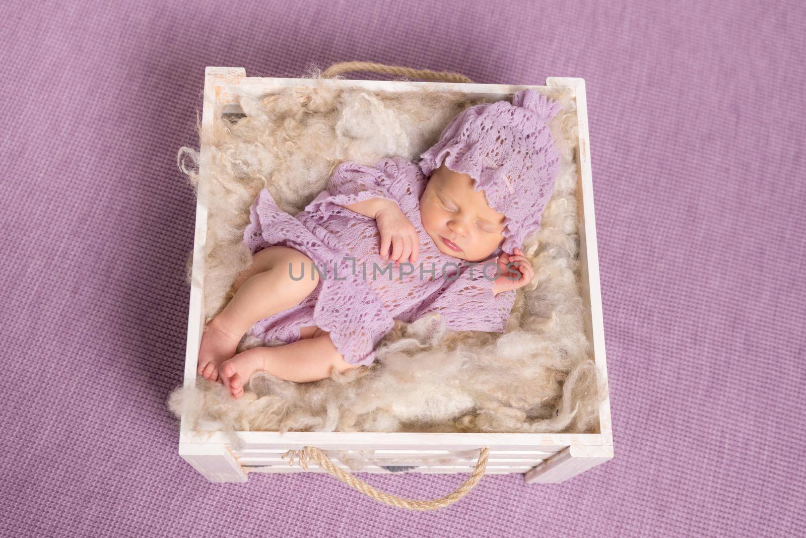 sweet newborn girl sleeping in square cot on violet background and cute hat with costume