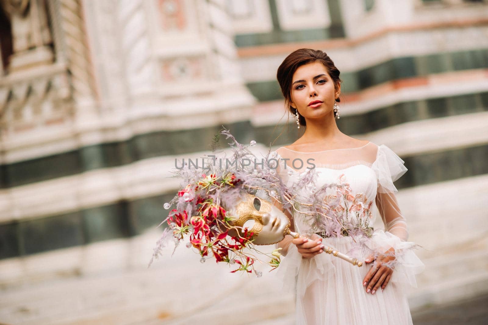 the girl-bride is with beautiful flower pattern as a mask in Florence, stylish bride in a wedding dress standing with a mask in the Old town of Florence. Model girl in Florence.