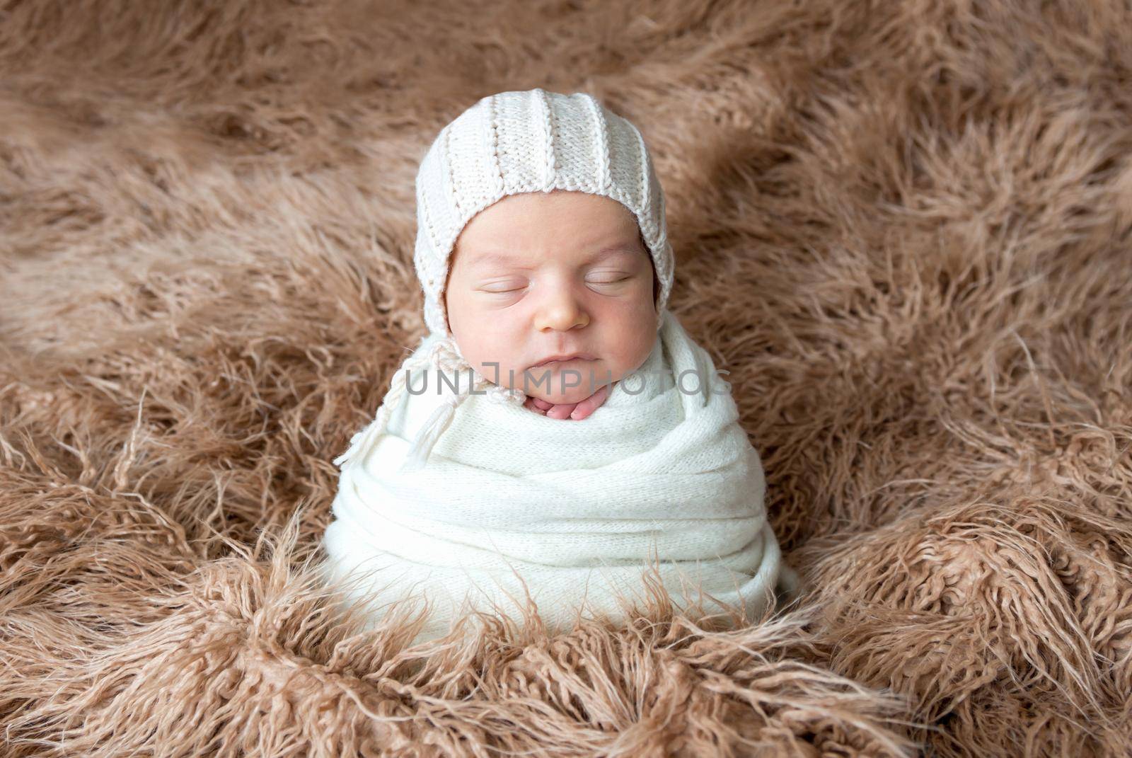Newborn child swaddled in a white woolen cloth is sleeping on brown fluffy plaid