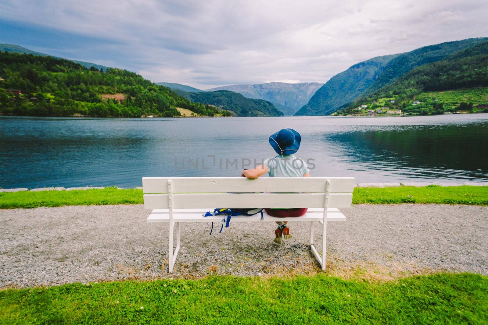 Woman sitting on a bench looking at the fjord in Ulvik, Norway. Fjord coastal promenade in Ulvik, Hordaland county, Norge. Lonely tourist in hat sits back on bench and admires scenery Scandinavia.