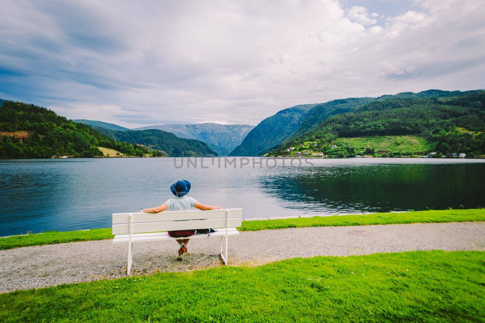 Woman sitting on a bench looking at the fjord in Ulvik, Norway. Fjord coastal promenade in Ulvik, Hordaland county, Norge. Lonely tourist in hat sits back on bench and admires scenery Scandinavia by Tomashevska