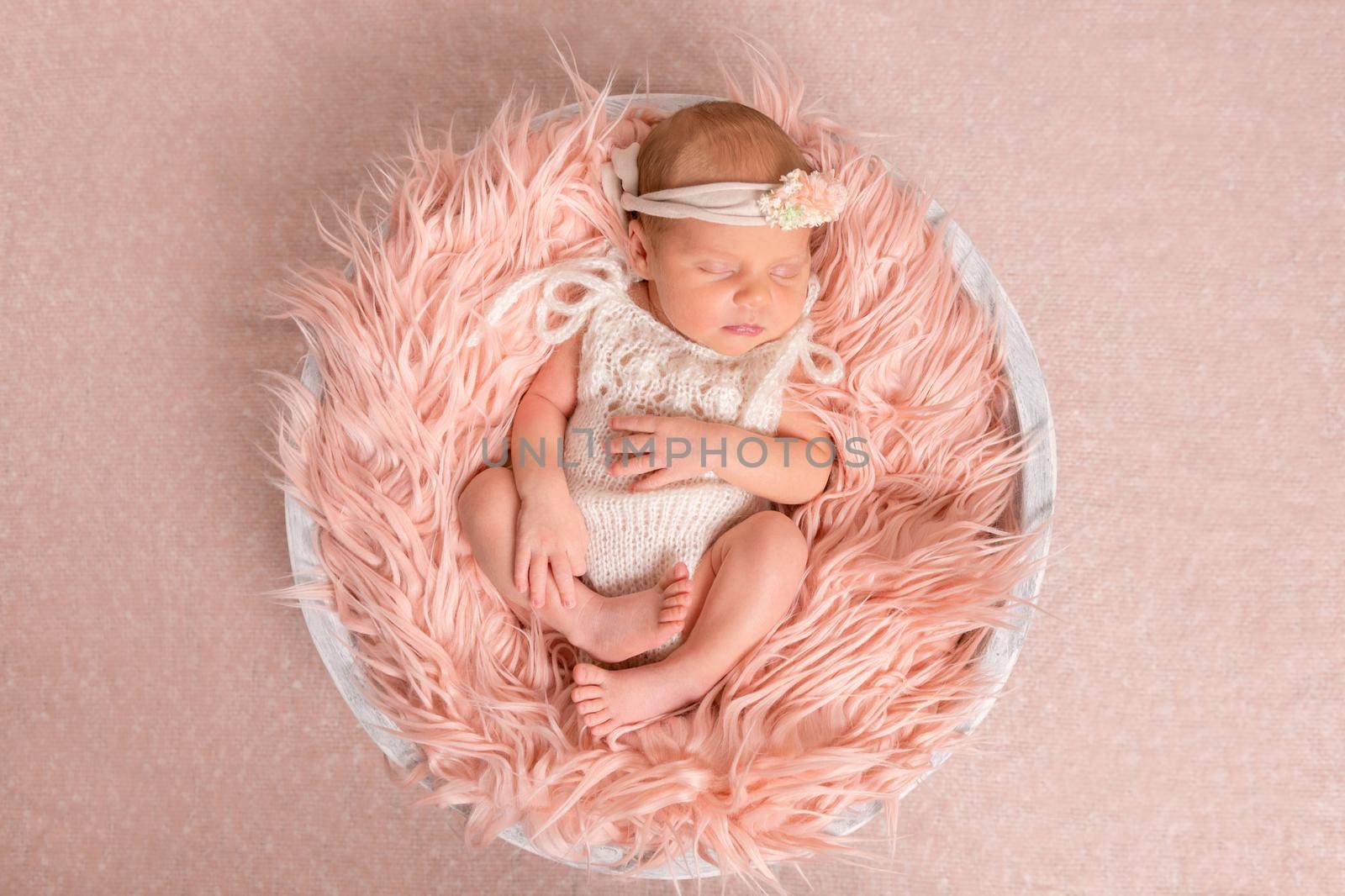 sweet newborn girl in white romper sleeping on round cot with fluffy blanket, top view
