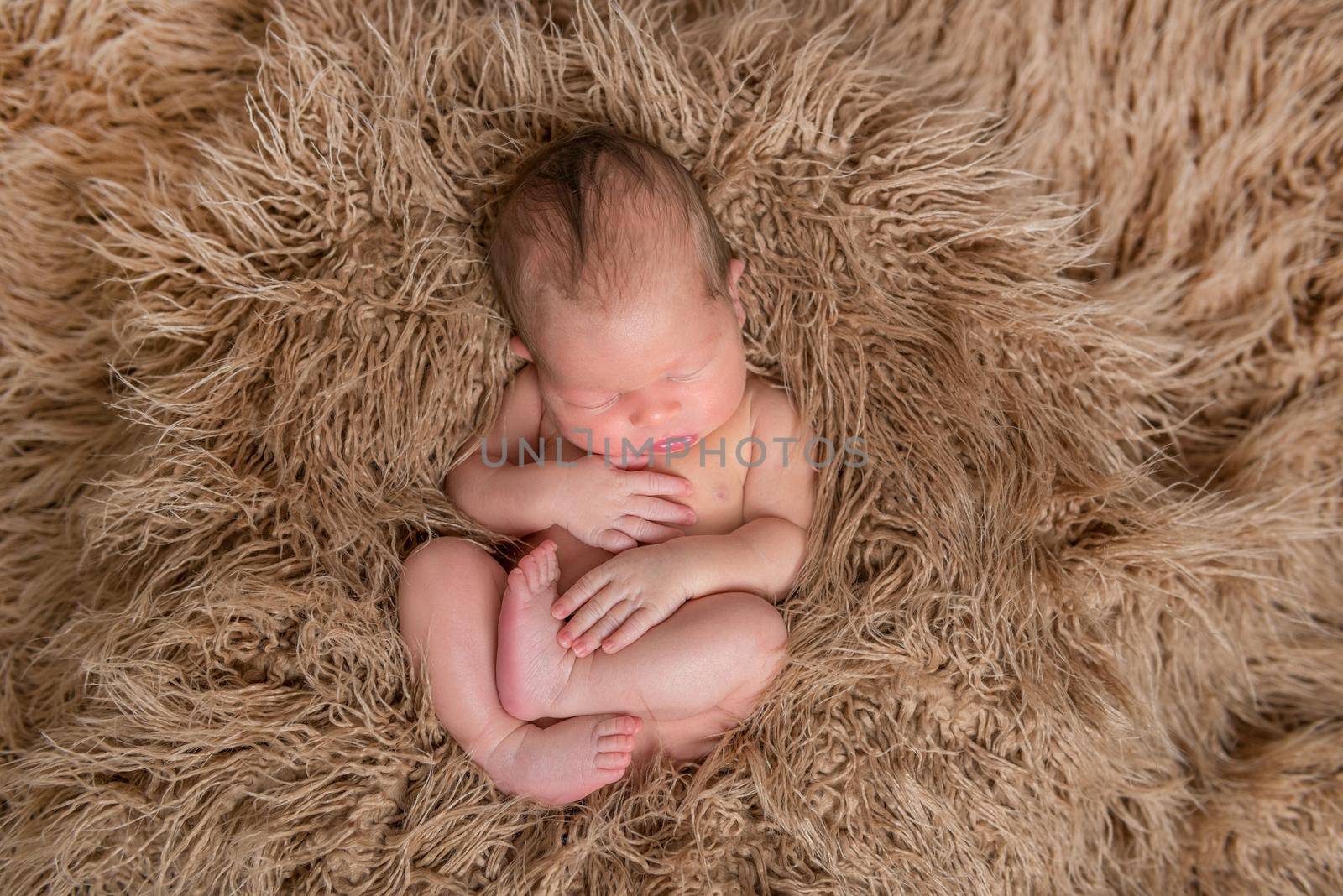 Naked sleeping baby curled up on a soft furry brown pillow, topview