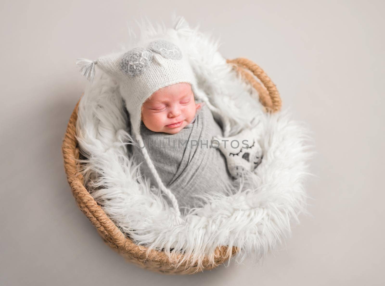 Little cute baby in white knitted beanie covered with gray coverlet sweetly sleeping on white soft blanket with toy owl in the basket