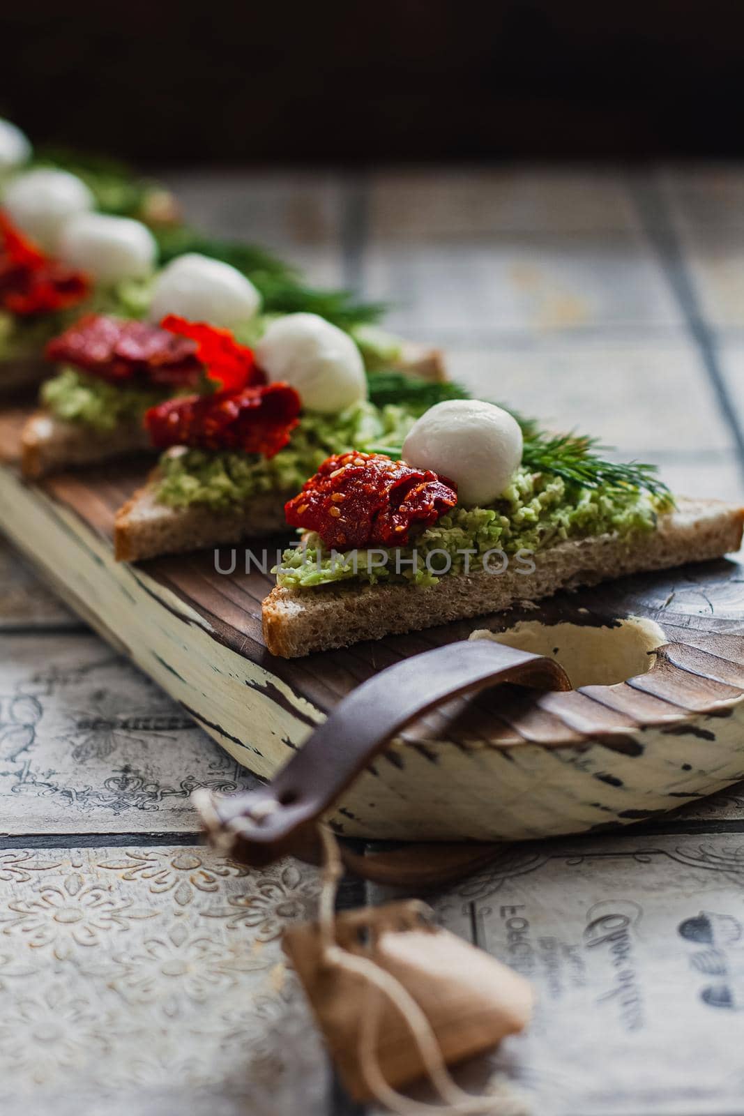 Mini triangle canapes bruschetta with avocado, mozarella and dried tomatos on the rustic board on the vintage tiled background.