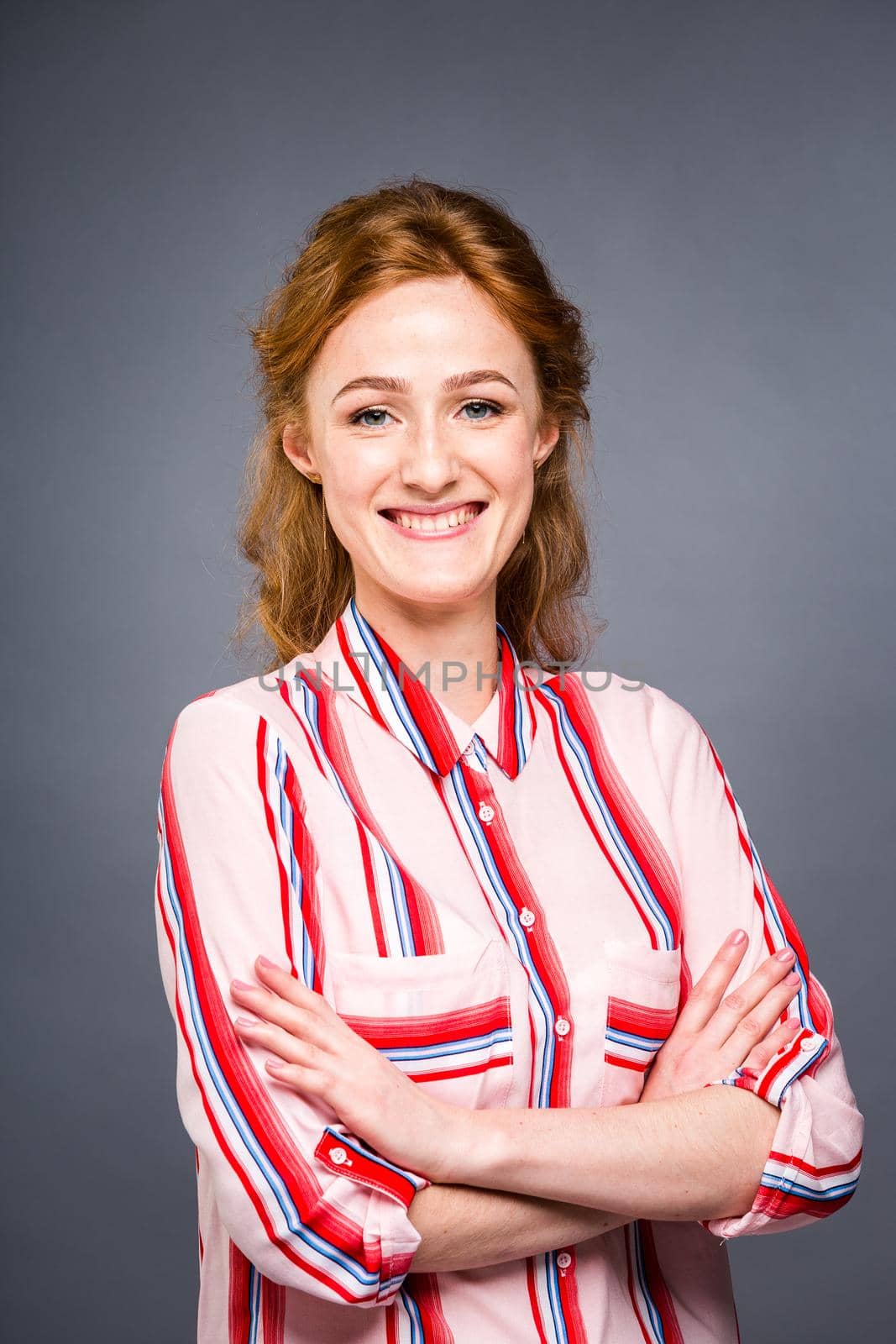 portrait of a young red-haired beautiful girl in the studio on a gray isolated background. A woman stands and smiles in a red shirt with a short sleeve. Business concept.